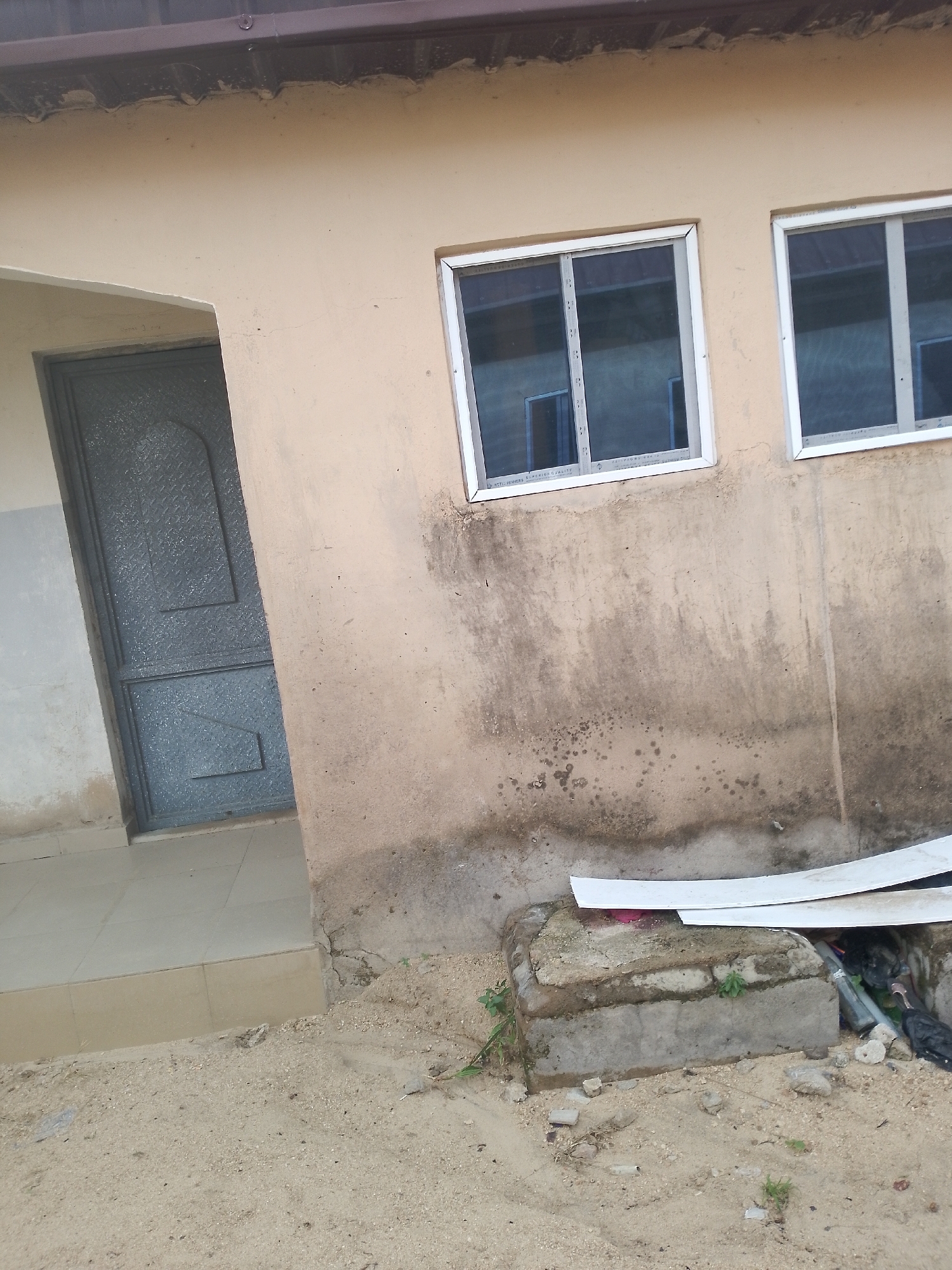 The image shows the exterior of a building with a light brown, stucco-like wall. To the left, a dark gray, textured door is visible, set within an opening. The door appears to be made of metal. Next to the door is a tiled walkway or step. Above the door and extending across the wall are two windows with white frames. The wall has some discoloration and dirt marks, particularly towards the bottom. In the foreground, the ground is covered in sand and small rocks, with some debris and a concrete structure. The roofline of the building is visible at the top of the image.