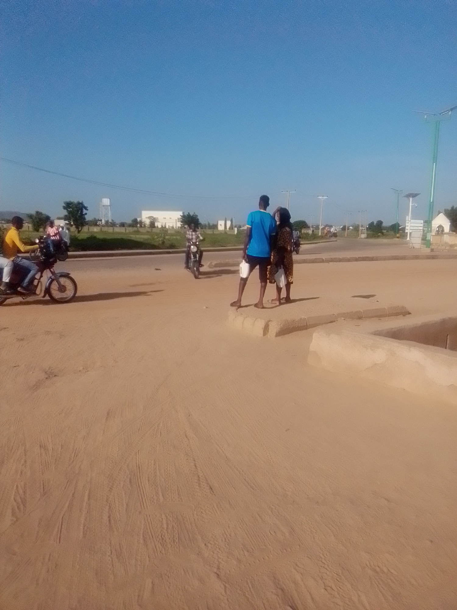 The image depicts a sunlit street scene in Kwadon, Nigeria, under a clear blue sky. In the foreground, a sandy road occupies the lower half of the frame, marked with tire tracks. On the right side of the road, two individuals are standing on a raised concrete platform. One person, wearing a blue t-shirt and dark shorts, holds a white container in their left hand. Beside them, another person, dressed in a patterned brown and black dress, also holds a white container.
Further down the road, two motorcycles are visible. On the left, a motorcycle is being ridden by a person wearing a yellow shirt and blue pants, with another person on the back wearing a striped red and white shirt. On the right side of the road, a motorcycle is being ridden by a person wearing a checkered shirt.
In the background, a paved road curves towards the right. Beyond the road, there is a low-lying landscape with trees, a white building, and a water tower. Several lampposts, some equipped with solar panels, are scattered throughout the area. The overall impression is a snapshot of everyday life in a Nigerian town.