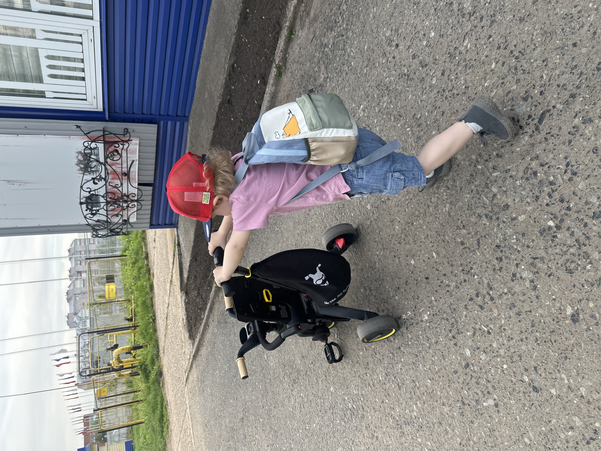 A child wearing a red cap, pink shirt, denim shorts, and grey shoes is riding a black tricycle on a paved surface. The child also has a small backpack on. To the left, there is a building with blue siding and a white window. In the background, there are yellow fences and flags, indicating that the location may be in Votkinsk, Russia. The flags may be referencing that city's or region's flag.