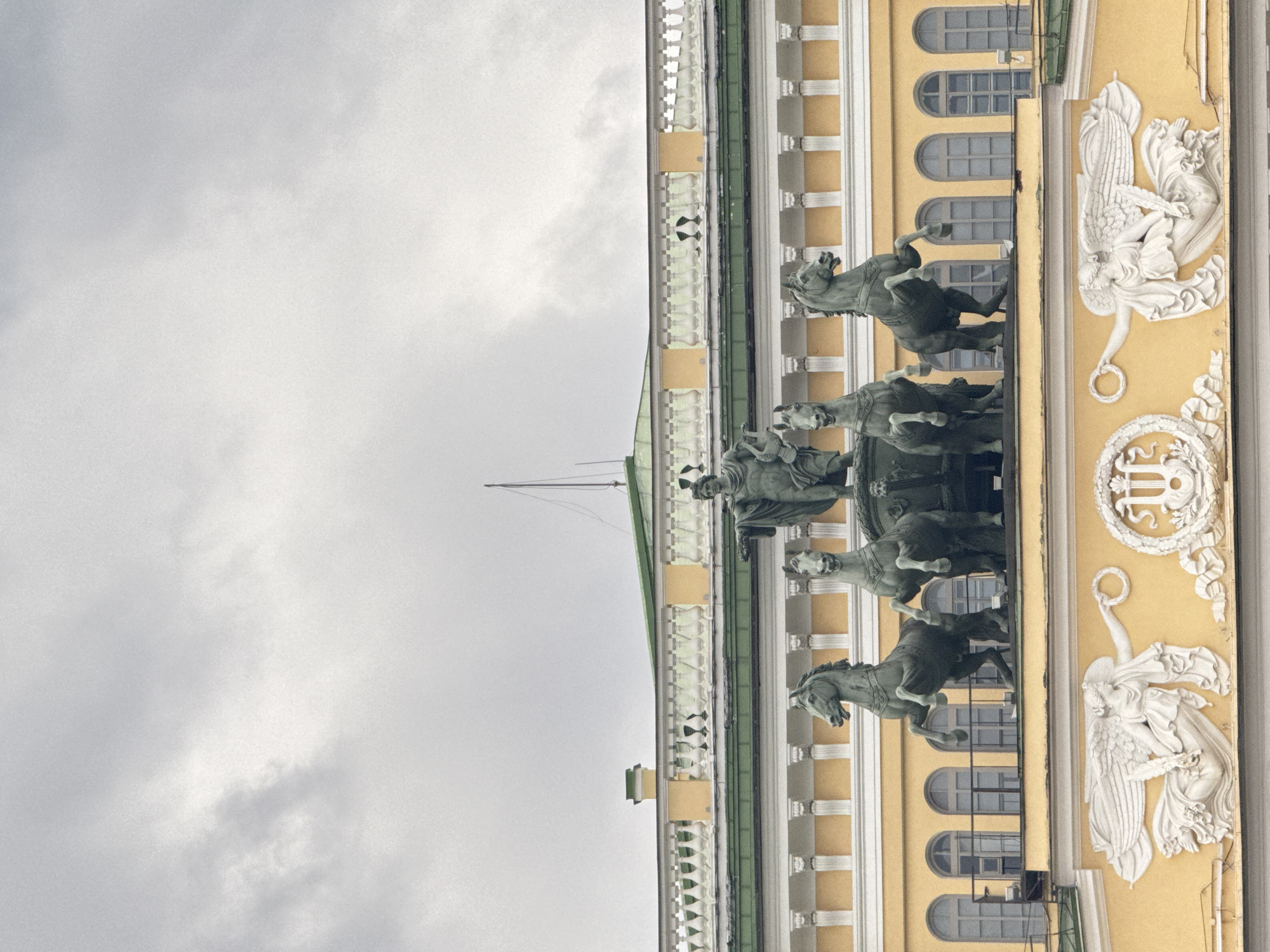 The image depicts a partial view of the General Staff Building in Saint Petersburg, Russia, specifically the triumphal arch section. On the building's facade are visible sculptural elements, including a quadriga (a chariot drawn by four horses) and decorative figures. The sky is overcast, suggesting a cloudy day. The color palette is dominated by yellow (facade), gray (sculptures and sky), and white (architectural details). The building appears to be well-maintained and of historical significance.