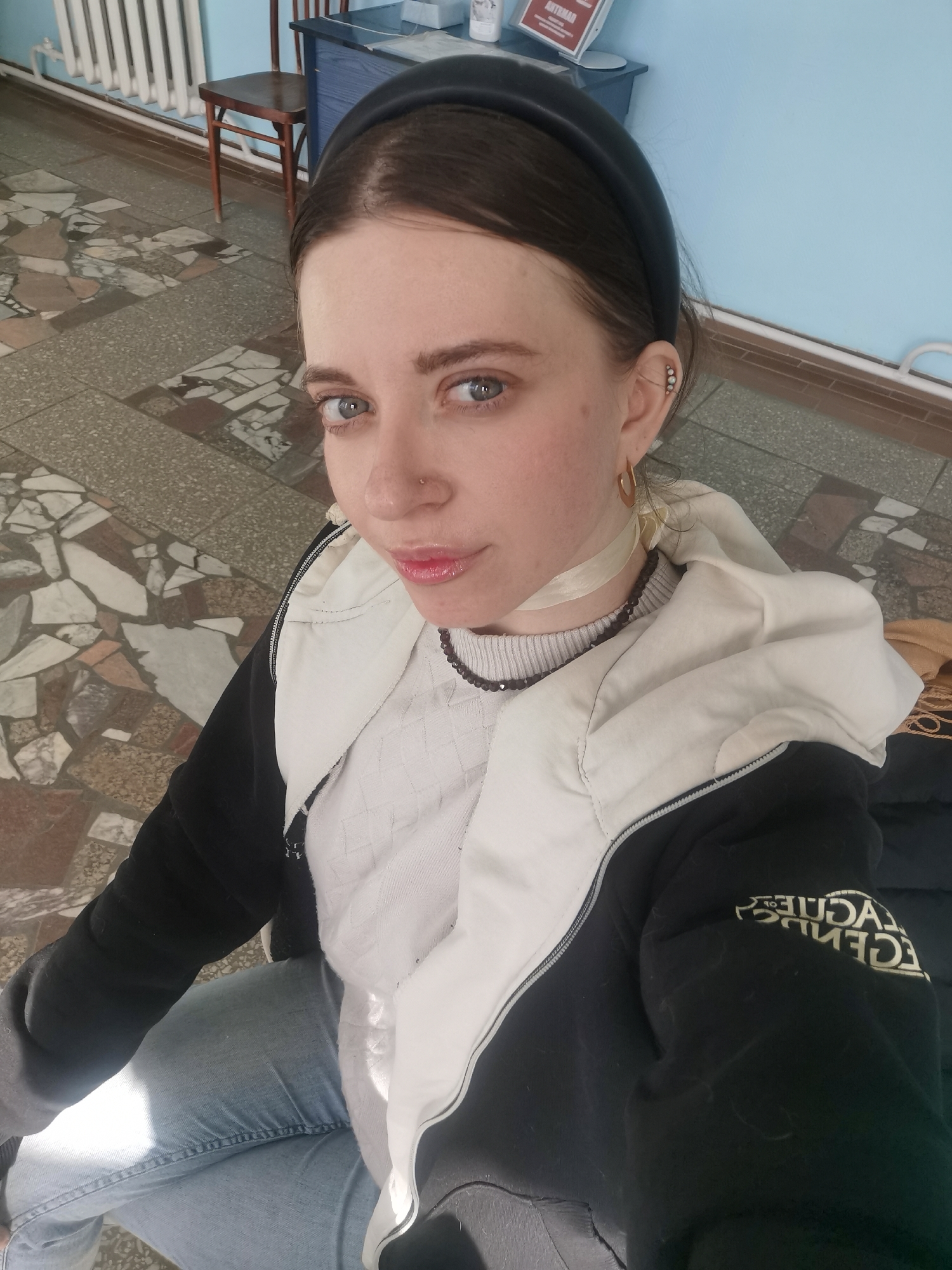 A young woman with dark hair and blue eyes is the primary subject of this high-angle selfie, looking directly into the camera with a neutral expression and slightly parted lips. She wears a black headband, a small nose stud, and multiple ear piercings, including a linear arrangement of small, light studs and a single hoop earring on her visible left ear. A light-colored ribbon or choker is tied around her neck, layered with a dark beaded necklace. Her clothing includes a white or cream textured sweater, a black and white puffer jacket, and light blue jeans. The black sleeve of her jacket bears the mirrored text "LEAGUE OF LEGENDS".
The setting appears to be an indoor public or institutional space, possibly a waiting area or hallway. Behind her is a light blue wall with a white horizontal radiator running along its base. The floor features a mosaic-like pattern composed of irregular pieces of gray, white, brown, and reddish-brown tiles. A small wooden stool with a brown seat and dark legs is visible to her left. Behind the stool, part of a dark blue desk or cabinet can be seen, on which a white sign with faint red Cyrillic text (possibly "АКТРИАЛ") is placed. The lighting suggests daytime, with natural light entering from an unseen source, creating soft shadows. The woman is likely seated or crouching, holding the camera above her.