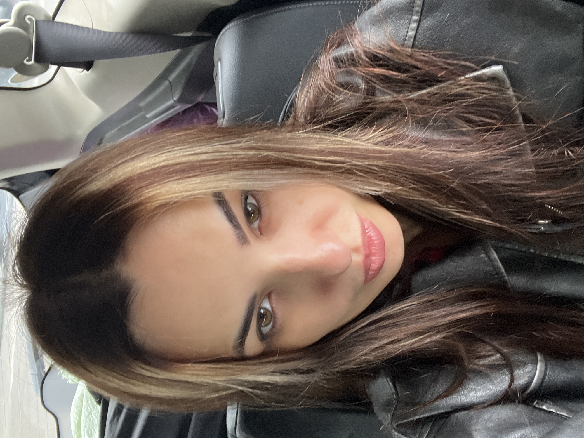 The image displays a close-up, headshot-style portrait of a female individual seated inside the rear passenger compartment of a vehicle. Her face is directed towards the camera, with a neutral expression and slightly parted lips, which have a light pink color. She possesses dark brown eyes and long, dark brown hair with distinct lighter, possibly blonde or light brown, highlights framing her face and distributed throughout her hair, which is parted down the middle. She is wearing a black leather jacket with visible zipper details on the front. The background reveals components of the car's interior, including a dark upholstered seat, a light-colored upper car trim, and a black seatbelt strap and anchor mechanism visible on the upper right side of the frame. A blurred light green fabric item is partially visible in the upper left background, and a purple object is present behind her head on the right. The location is inside a vehicle; no specific city or external geographic details are identifiable.