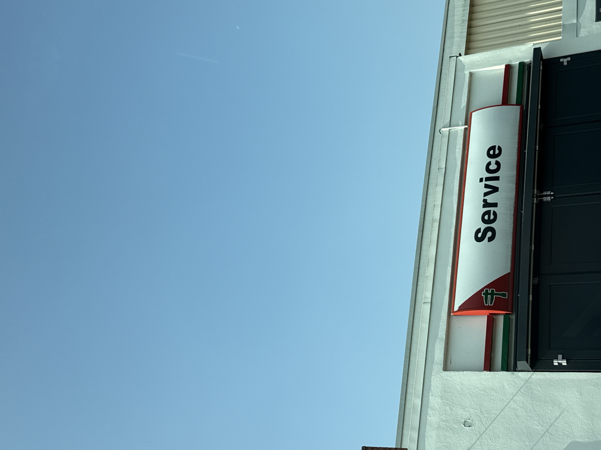 An outdoor, eye-level shot shows a building with a sign that says "Service" in black, bold lettering. The sign is on a white background with red and green stripes. To the left of the word "Service" is a red and green emblem. The building has a light-colored stucco exterior and a darker colored door below the sign. To the right of the building, a section of ribbed metal siding is visible. The sky above is clear and blue. The image was taken in Bad Sachsa, Germany.