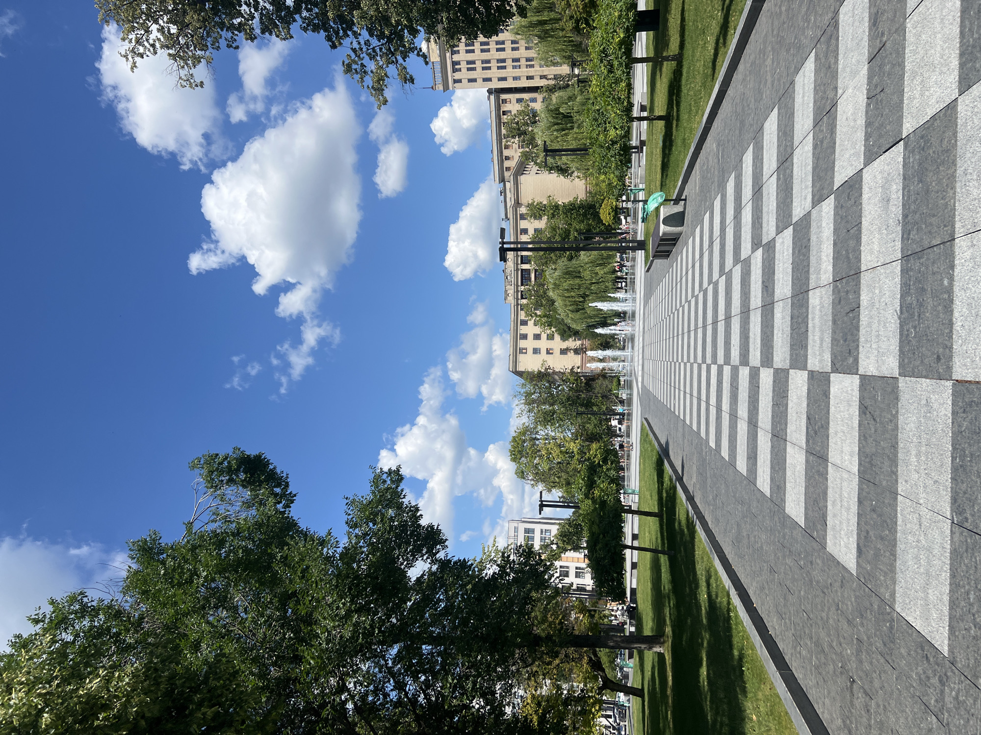 This is a photograph of a public park or plaza in Kharkiv, Ukraine, featuring a checkered pavement walkway leading towards a series of fountains in the background. The walkway is made of alternating dark grey and light grey stone tiles. Lush green grass borders the walkway, with several trees and manicured bushes scattered throughout the park. In the mid-ground, several people are visible walking and sitting, enjoying the outdoor space. Behind the park, a multi-story building complex with a light-colored facade and numerous windows can be seen. The sky above is a vibrant blue, dotted with fluffy white clouds, suggesting fair weather. The overall scene depicts a serene and well-maintained urban public space.