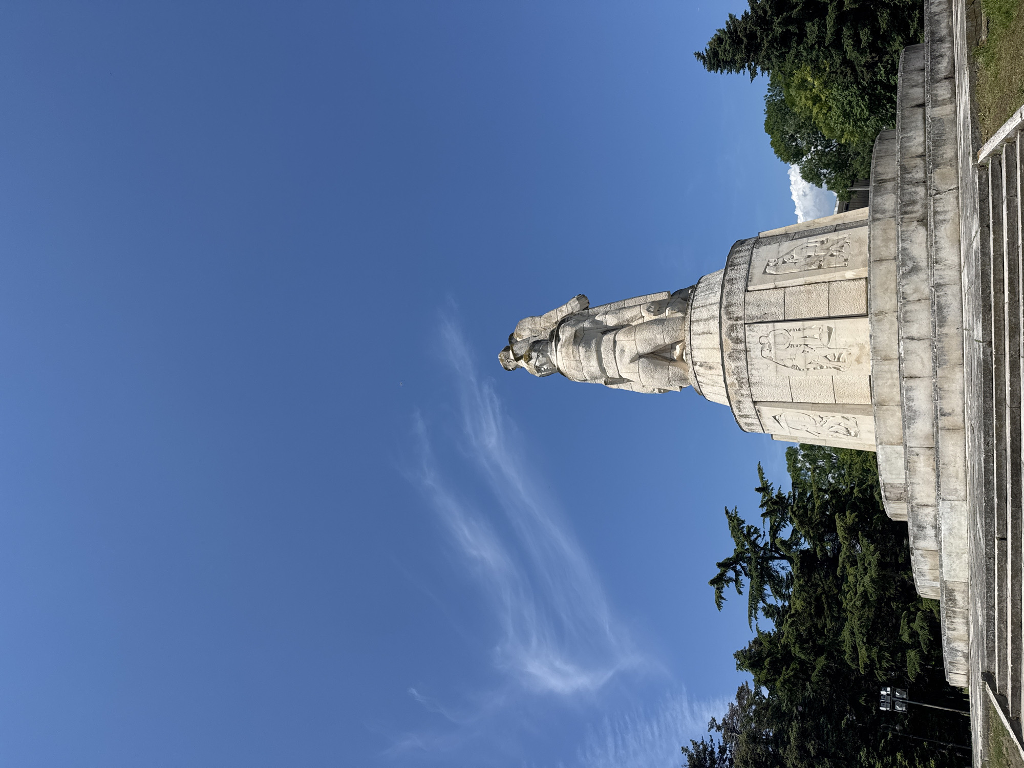 The image captures a towering stone monument or statue, presented from a low, upward-looking angle that imparts a significant tilt within the vertical frame. The monument is constructed from light grey, weathered stone, featuring multiple sculpted figures on its upper part and a cylindrical base adorned with intricate relief carvings depicting various scenes.
To the right of the monument, a set of wide, grey stone steps leads upwards, matching the material and style of the main structure. The monument and steps are framed by lush green trees, including both tall coniferous types and broader deciduous trees, suggesting a setting within a park or memorial grounds.
The sky is a brilliant, clear blue, indicative of a bright, sunny day. A few delicate, wispy white clouds are visible, particularly trailing across the upper left portion of the sky. A tiny, distant dark speck is also discernible high in the sky. No specific human activity is taking place, and no discernible text is visible on the monument or elsewhere in the scene. The overall impression is one of solemnity and historical remembrance.