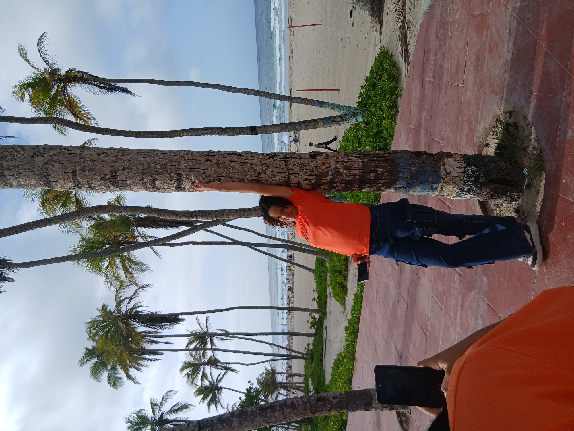 The image shows a woman posing by a palm tree on Playa El Agua, Venezuela. She is wearing an orange t-shirt and blue cargo pants, with her left arm extended upwards and leaning against the tree. She is holding a cell phone in her right hand. Behind her, the beach is visible with rows of beach umbrellas and people relaxing. The ocean is calm with small waves. The foreground has a red tiled walkway with a palm tree in the center. Another person is partially visible in the lower left corner, holding up a phone, possibly taking a picture of the woman. The sky is partly cloudy.