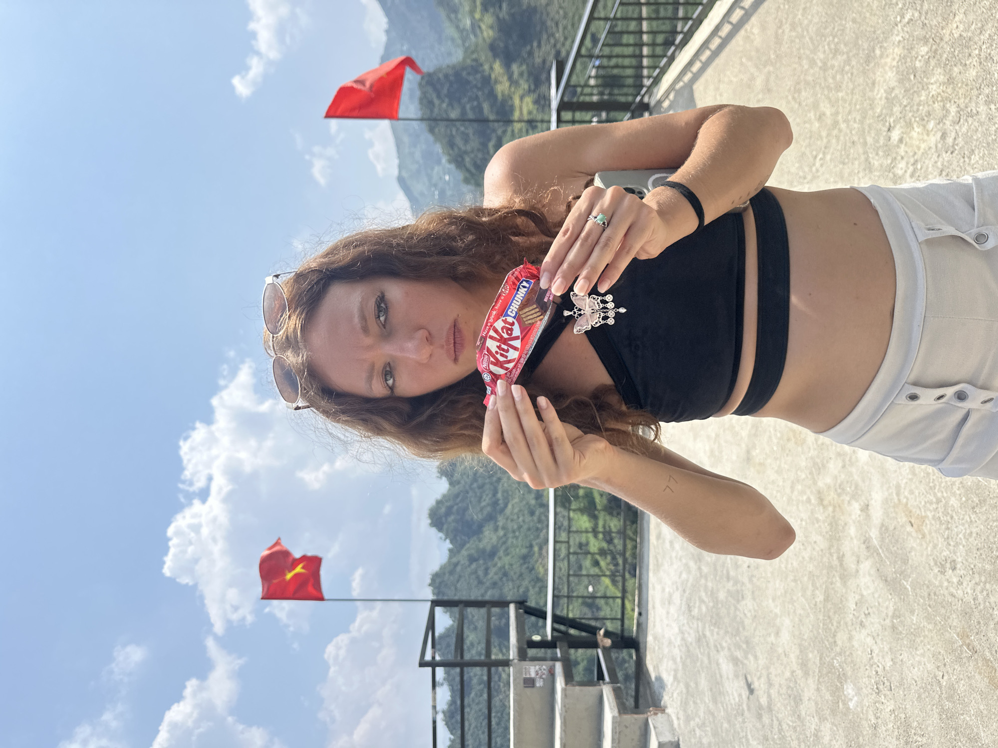 A young woman stands on a concrete surface in Lũng Phìn Commune, Vietnam, against a backdrop of mountains and a partly cloudy sky. She is holding a Kit Kat Chunky candy bar. Two red flags are visible.