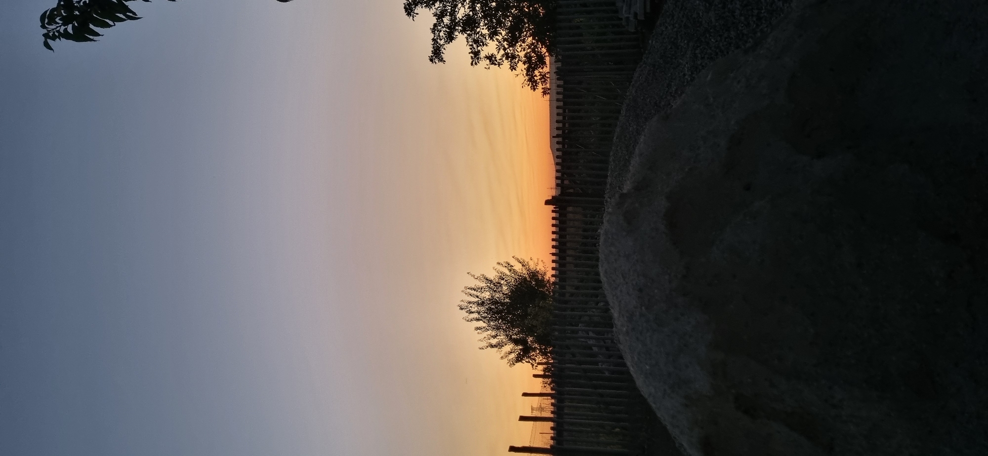 The image captures a twilight scene in Blizhnee, Ukraine. In the foreground, a large, dark pile of aggregate, possibly sand or gravel, dominates the lower portion of the frame. Behind it, a rustic wooden fence with vertical posts runs horizontally across the mid-ground. Beyond the fence, silhouetted trees with dense foliage are visible against the horizon. The sky displays a gradient of colors, transitioning from a soft blue at the top to warm orange hues near the horizon, indicating sunset or sunrise. The overall environment appears to be an outdoor space, possibly a construction site or a rural property, characterized by earthy materials and natural elements.