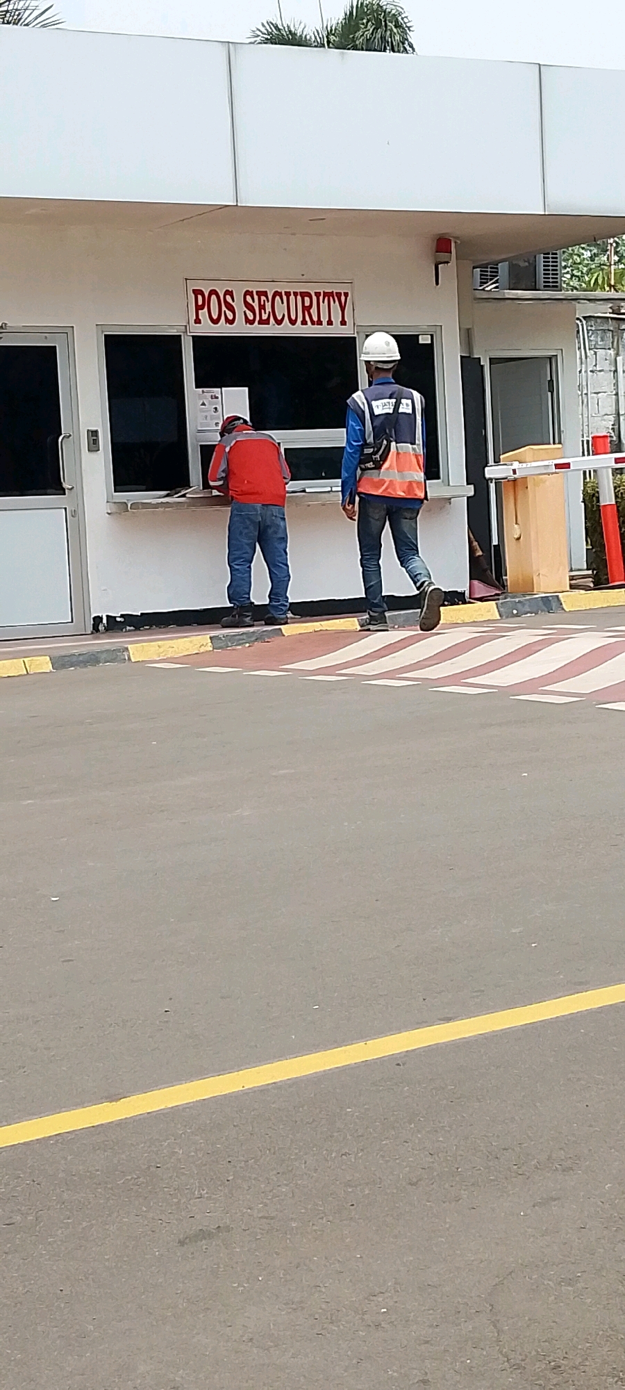 The image shows a security post labeled "POS SECURITY" located in Depok, Indonesia. Two individuals are present. One person, wearing a red jacket and blue jeans, is standing at the window of the security post, appearing to interact with someone inside or handle paperwork. The second person, wearing a white hard hat, a blue long-sleeved shirt, and an orange high-visibility vest over it, is standing outside the security post and walking away. A barrier gate is visible to the right of the security post, with red and white striped bollards and a white pole. The ground in front of the security post is paved with asphalt, marked with yellow and red-white striped lines, suggesting a pedestrian crossing or traffic demarcation. Palm trees are visible in the background, indicative of a tropical climate. The security post itself is a white building with large windows.