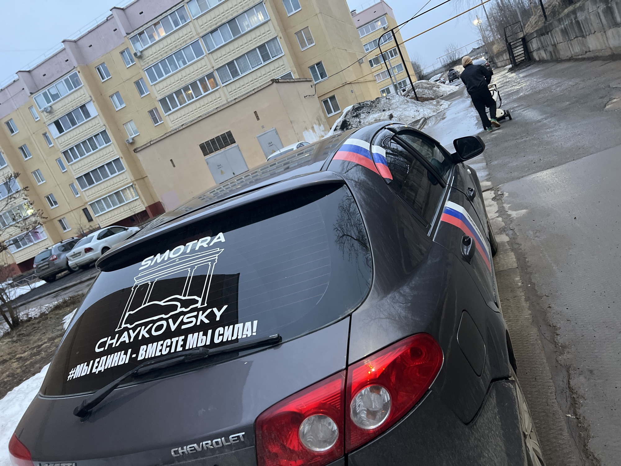 A gray Chevrolet car is parked on a street next to a sidewalk in what appears to be a residential area. The car has Russian flag decals on the rear side panels and a large sticker on the rear window featuring the word "SMOTRA" above a drawing resembling a colonnade, followed by "CHAYKOVSKY" and the phrase "#МЫ ЕДИНЫ - ВМЕСТЕ МЫ СИЛА!" (We are united - together we are strong!).
In the background is a multi-story apartment building with yellow walls and numerous windows. Patches of snow are visible on the ground near the car and the building. A person dressed in dark clothing is walking away from the camera, pushing a stroller on the sidewalk. The street is wet, indicating recent precipitation or melting snow. The general environment suggests it might be a cold or overcast day.
Based on the information on the rear window, the city is likely Chaykovsky, Russia, though the image was taken in the city of Noviy.