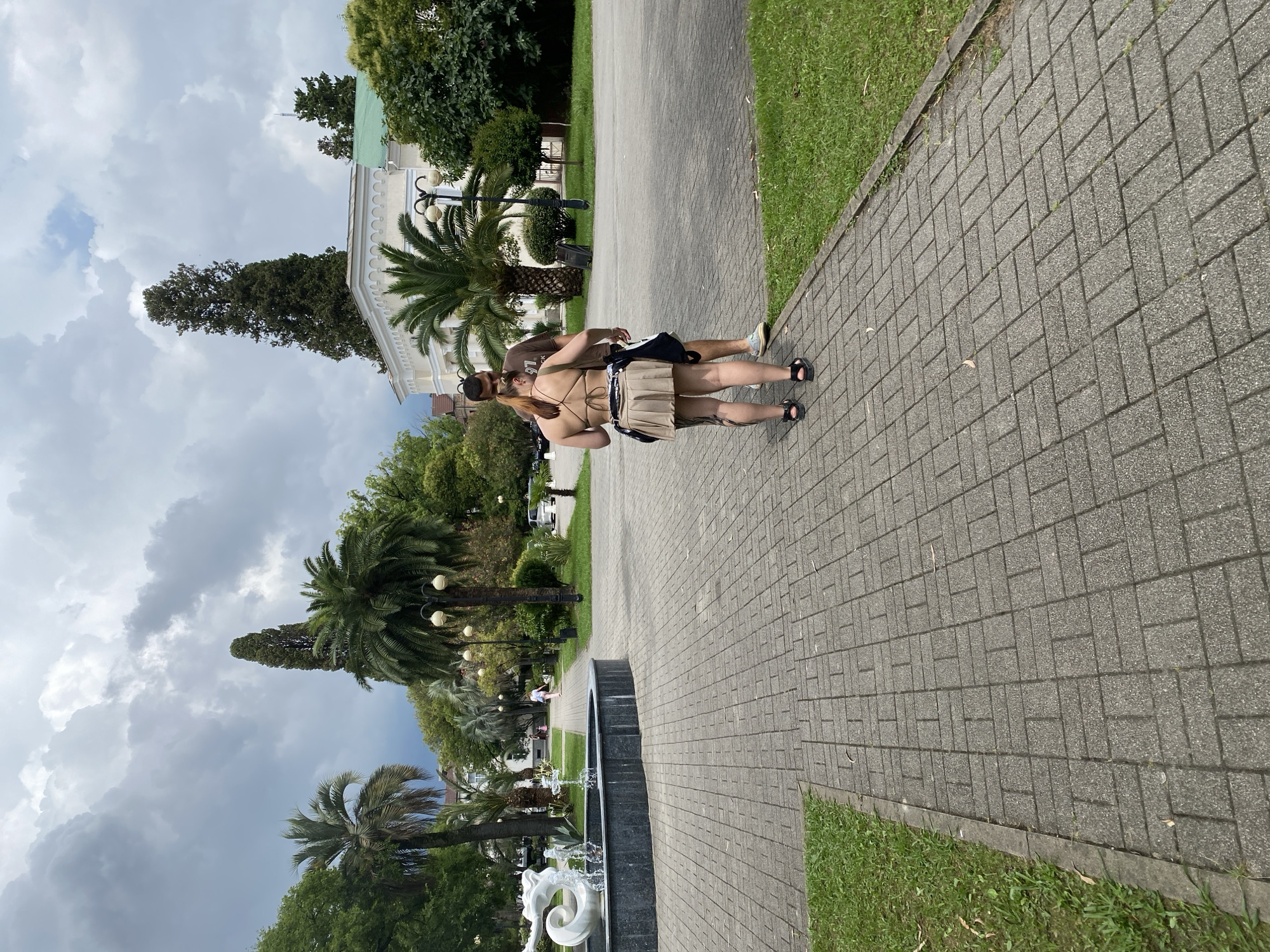 In Sokhumi, Abkhazia, two individuals, a man and a woman, stand on a paved walkway in a park on a cloudy day. The woman, facing away from the camera, wears a tan bikini top and a pleated tan skirt. The man, partially obscured by the woman, wears a brown t-shirt. Behind them, a circular fountain with a sculpture of a woman and water jets is visible to the left. The walkway is bordered by green grass. Lush trees, including palm trees and a tall cypress, surround the area. In the background, a white building with an arched facade and a green roof can be seen, along with streetlights and parked cars.