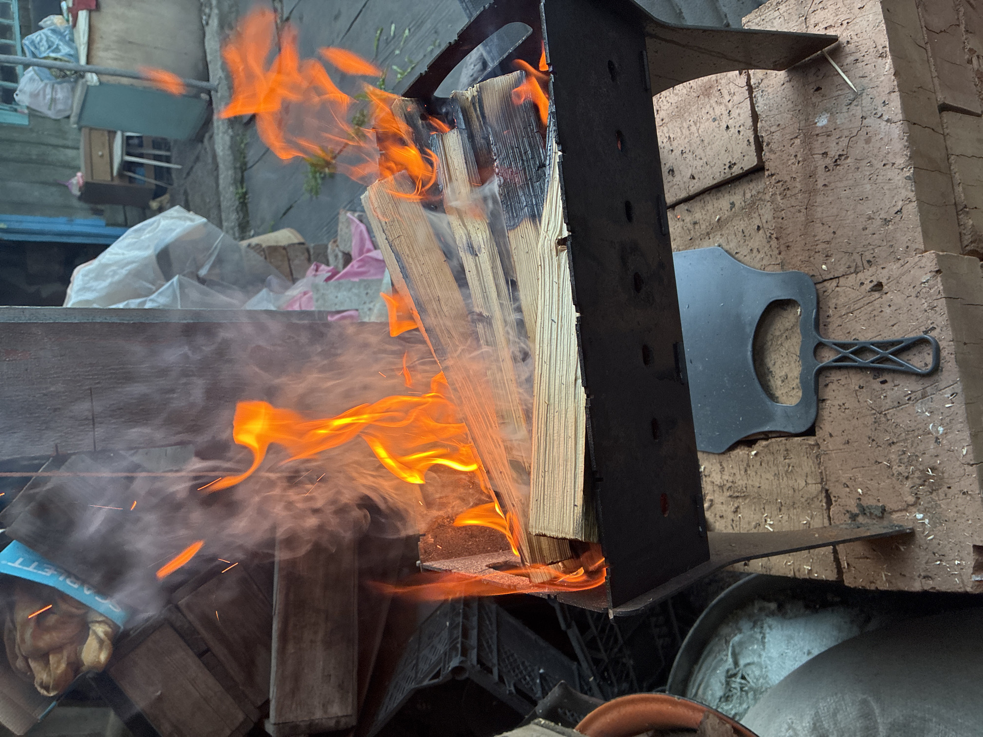 Here is a forensic description of the image:
The image shows a fire burning in a metal structure, which contains several pieces of wood stacked together. The flames are orange and yellow, and there is a significant amount of smoke. To the right of the structure is a brick wall and a black metal utensil. The scene is located outdoors in an environment containing miscellaneous objects, suggesting a backyard or similar space. A blue and white item appears near the fire. The location is listed as Unknown, Unknown.