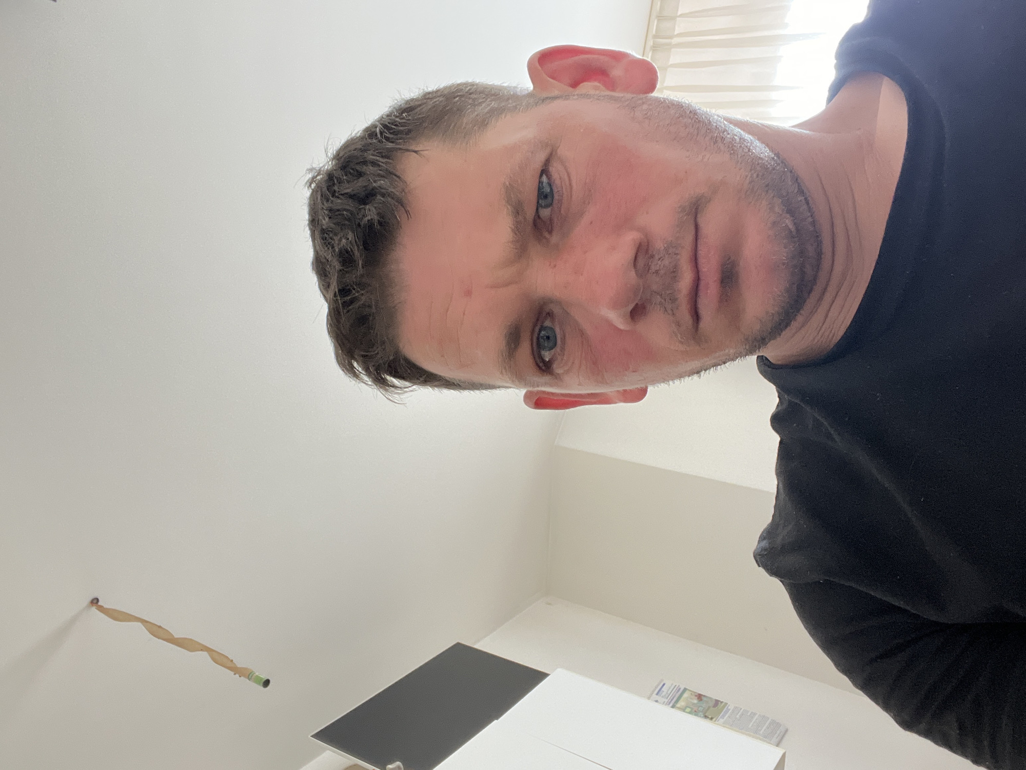 The image is a selfie taken indoors, likely in an apartment. The subject is a white male with short, dark hair, blue eyes, and a fair complexion. He is wearing a black t-shirt. His face is slightly flushed, and he has a concerned or questioning expression.
In the background, a white ceiling is visible, with a twisted paper decoration hanging from it, attached to a small green object at the bottom. To the left, there are two white wall-mounted units, one with a dark gray rectangular panel on top, and a white one below with some papers attached. The right side of the image shows a cream-colored curtain.
The specific location in Senago, Italy, cannot be determined from the visual information provided in the image.