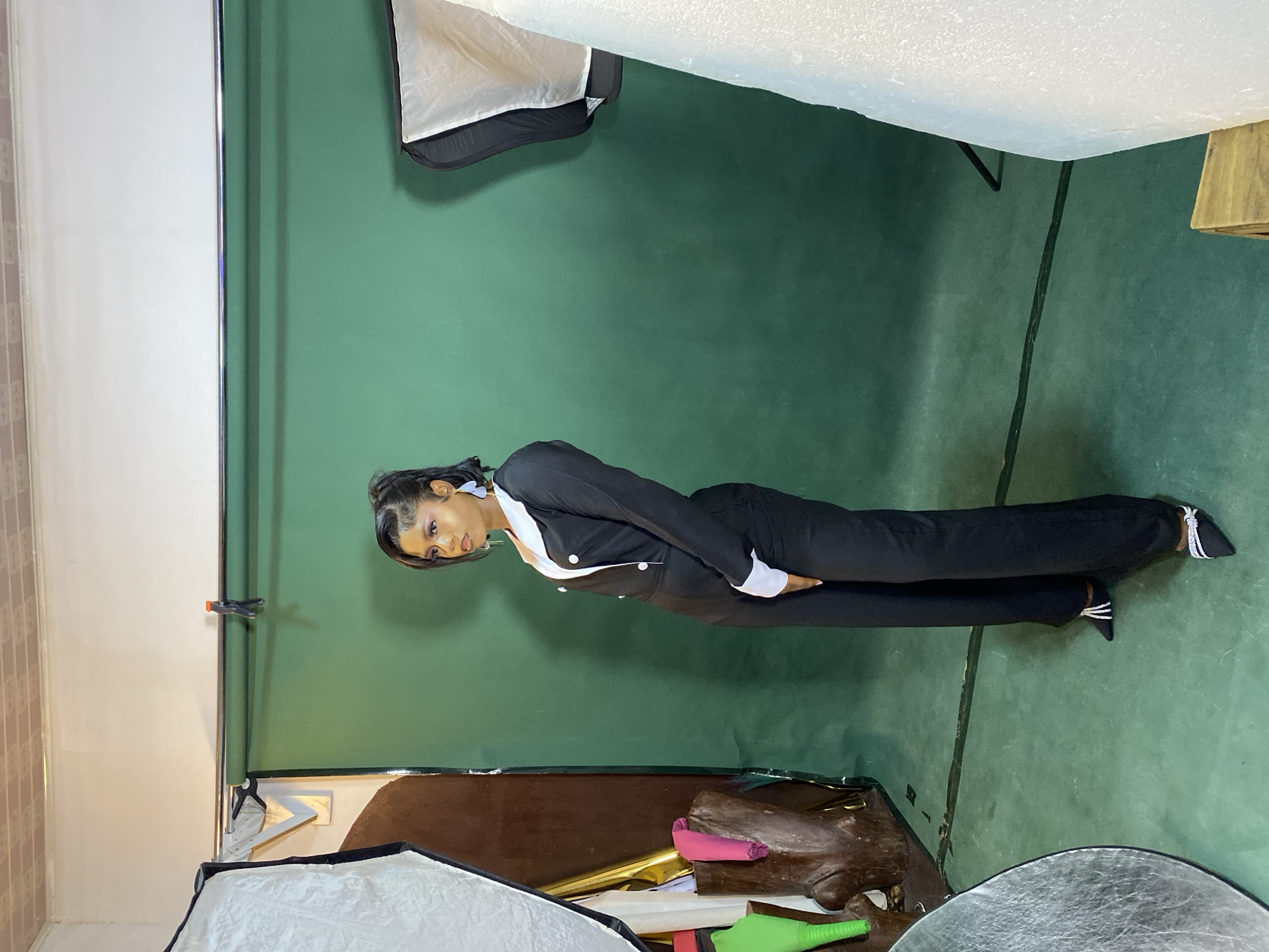 The image depicts a woman posing in what appears to be a photography studio in Jos, Nigeria. She is wearing a black pantsuit with white trim and pointed heels. The background is a green screen. Lighting equipment, including a softbox and reflector, are visible in the foreground. The walls of the studio are painted white.