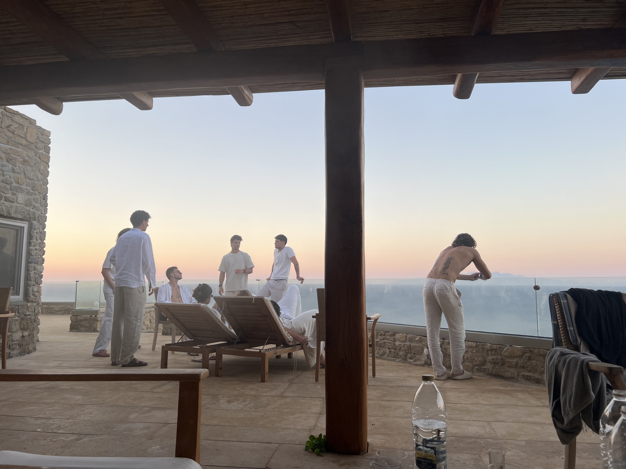 At sunset on a patio overlooking the sea in Unknown, Greece, a group of young men are gathered. The sky is a gradient of orange and blue, transitioning into twilight. The patio is constructed of stone and wood, with a covered area supported by wooden beams. In the foreground, a wooden table with chairs is visible, with a bottle of water and glasses on it. Several men are standing and lounging on chairs, dressed in light-colored clothing. One man, shirtless, stands with his back to the camera, looking out at the sea. Another man, wearing a white shirt and pants, stands near the center of the frame. The scene appears relaxed and social, with the men enjoying the view and each other's company.