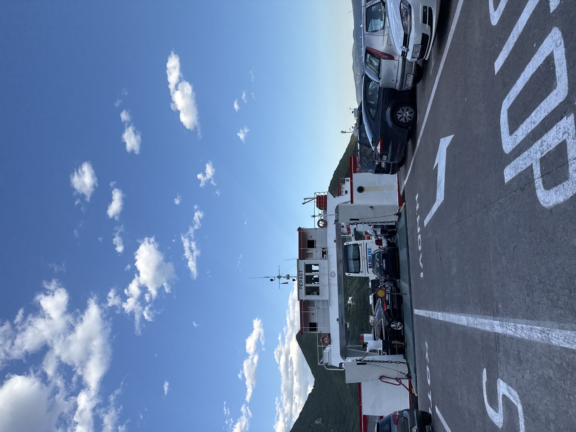 A ferry named PERAST is docked with several cars on its deck. A blue and white bus with "BLUE LINE" written on its side is also on the ferry. The ferry is in the waters near the city of Đurići, Montenegro, with green hills in the background under a bright blue sky with scattered white clouds. The deck of the ferry has white painted markings, including arrows indicating direction and the word "STOP" in large white letters. People are visible on the ferry, some standing near the vehicles. The scene appears to be a sunny day with good visibility.