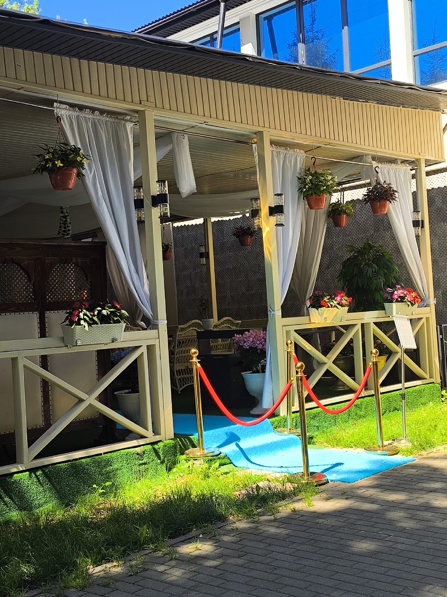 The image depicts an outdoor seating area, likely a cafe or restaurant, with a covered patio decorated with white drapes and hanging potted plants. A red velvet rope barrier is set up in front of a blue carpet, suggesting a VIP or entrance area. The patio is furnished with tables and chairs, visible through the drapes. The surrounding environment includes green artificial turf and natural grass, with a paved walkway leading towards the seating area. The architecture features wooden structures and a modern building with reflective blue windows in the background. The scene is illuminated by natural sunlight, casting shadows on the ground.