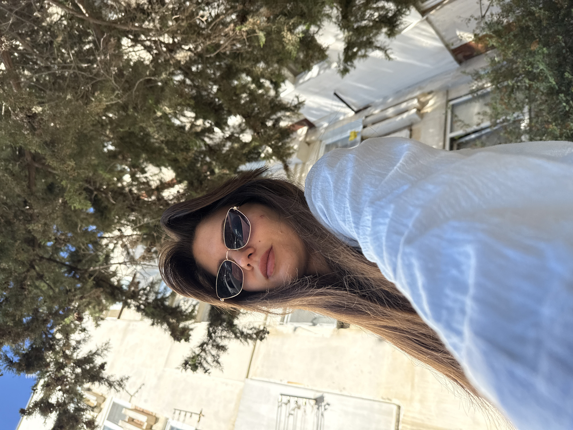 In an outdoor urban setting, a young woman with long, brown hair and sunglasses is captured from a low angle. She is wearing a white, loose-fitting shirt. The background features a building with windows and a tree with dense foliage. The sunlight casts shadows, indicating it is daytime. The woman is looking towards the camera, and her face is partially obscured by her hair and sunglasses. Her right arm is extended, suggesting she is holding the camera to take a selfie. The overall scene conveys a casual and relaxed atmosphere.