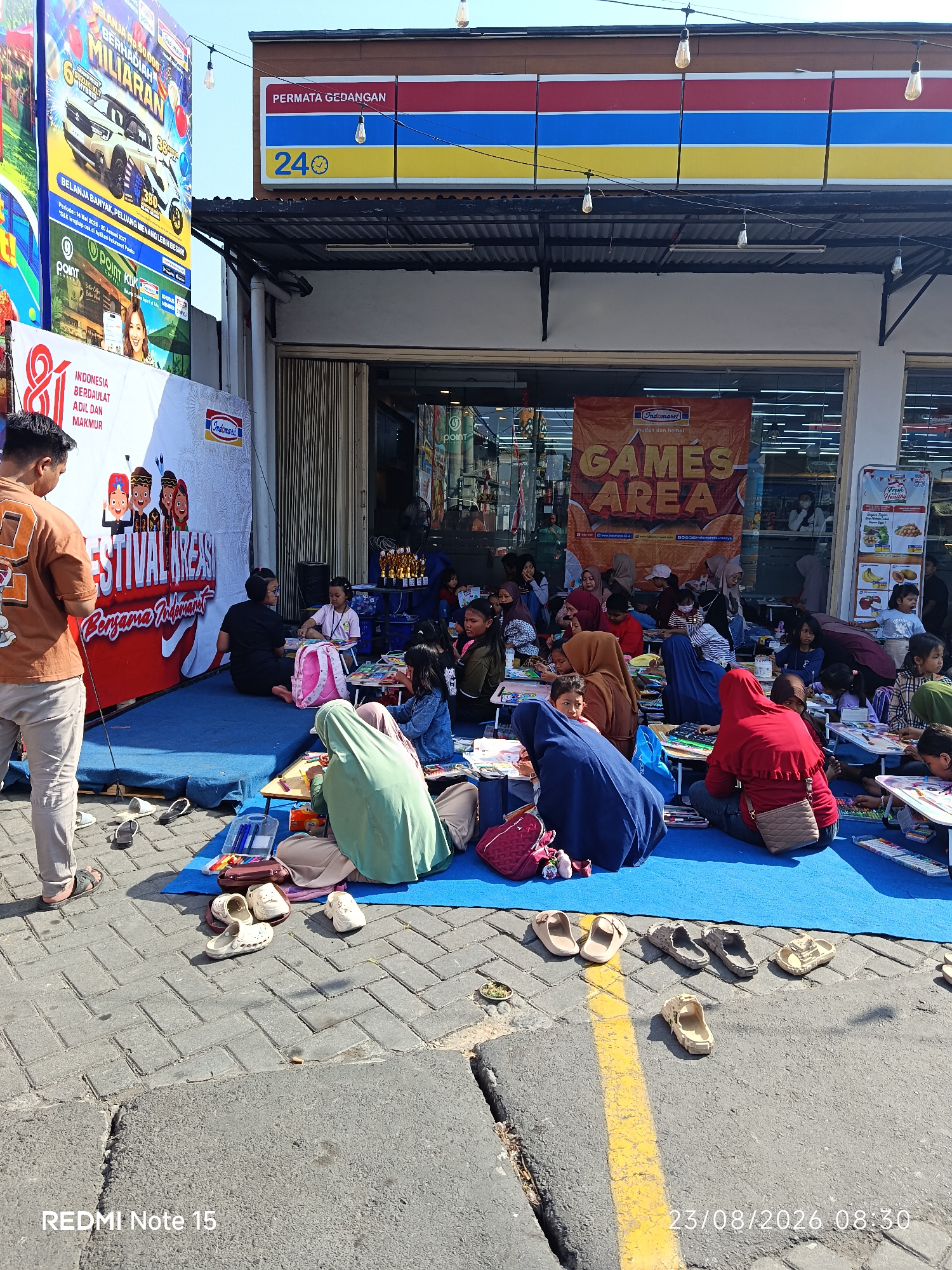 The image depicts an outdoor event in front of an Indomaret store in Keboansikep, Indonesia. A group of children and adults are seated on blue mats on a paved area, engaged in an activity, likely drawing or crafts, as indicated by the presence of art supplies like crayons and papers spread out on small tables. Several people, mostly women and children, are wearing headscarves. A large banner behind them reads "FESTIVAL KREASI Bersama Indomaret" (Indomaret's Festival of Creativity), suggesting a promotional or community event hosted by the store. Another banner to the right advertises "GAMES AREA". In the background, the Indomaret store's facade is visible, with its characteristic red, yellow, and blue signage. The overall scene is illuminated by natural daylight, with some artificial lights hanging from wires above. The pavement shows a pattern of interlocking bricks. Shoes are scattered on the ground near where people are seated, indicating they have been removed. A man in an orange t-shirt and beige pants is standing on the left, facing away from the camera, and appears to be observing the event. The environment suggests a sunny day.