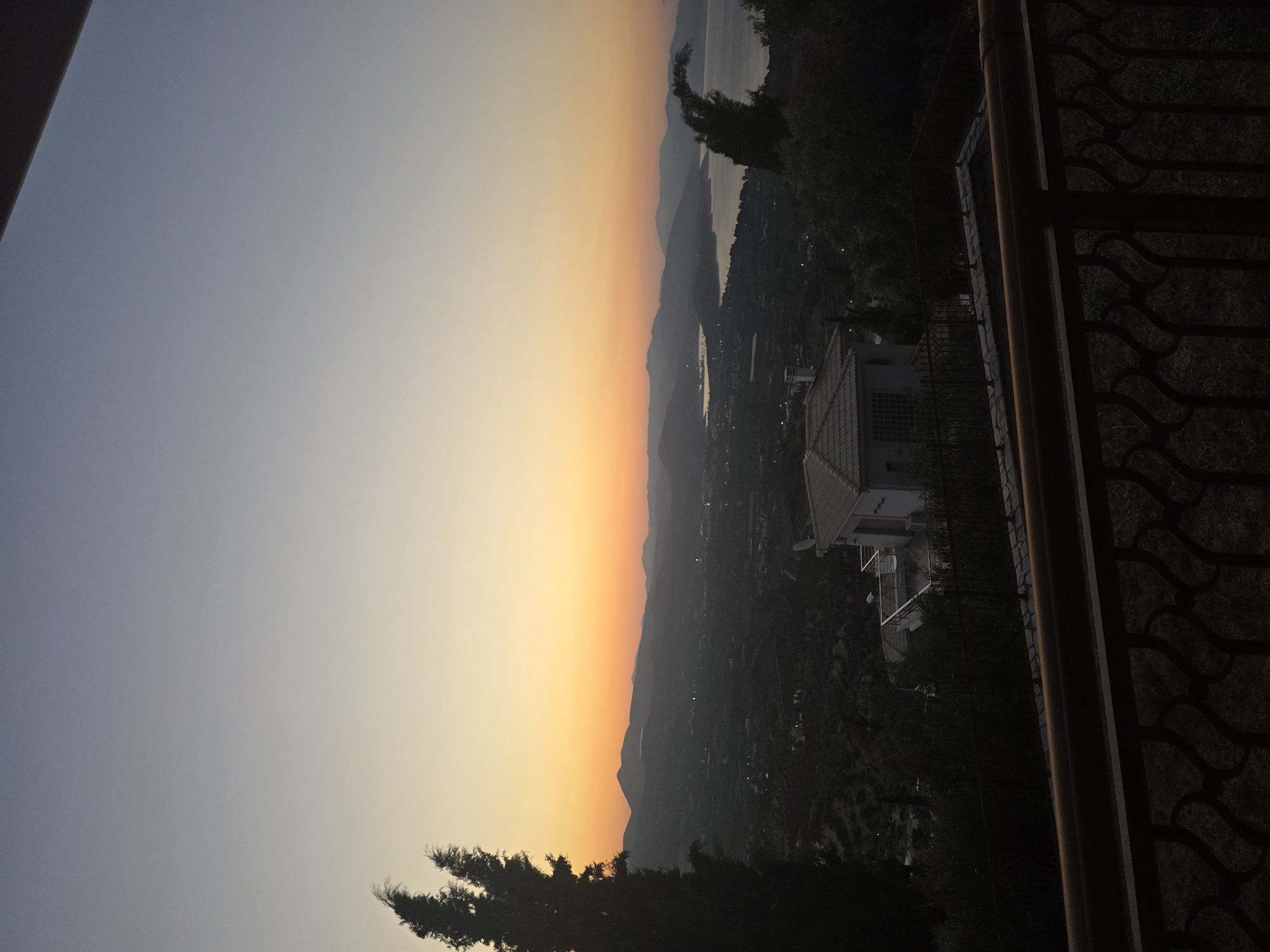 The image depicts a scenic view from a balcony overlooking a coastal landscape at dusk in the Municipal Unit of Asini, Greece. In the foreground, a dark, ornate metal railing runs horizontally across the bottom of the frame, with a glimpse of a paved walkway and greenery visible beyond it. A two-story house with a tiled roof and a balcony is partially visible on the right side of the frame.
The midground is dominated by a lush, green landscape dotted with trees and houses, suggesting a residential area. A prominent house with a white exterior and a light-colored tiled roof is situated in the middle ground, further back and slightly to the left of center. It features a balcony with furniture and a chimney.
The background showcases a dramatic sunset over a body of water, likely the sea, bordered by silhouetted hills and mountains. The sky transitions from a pale blue at the top to soft oranges and yellows near the horizon, indicative of the golden hour. The overall atmosphere is tranquil and serene. The time of day is clearly sunset, and the location is a residential area with views of the coast and surrounding hills. There are no people visible in the image.
