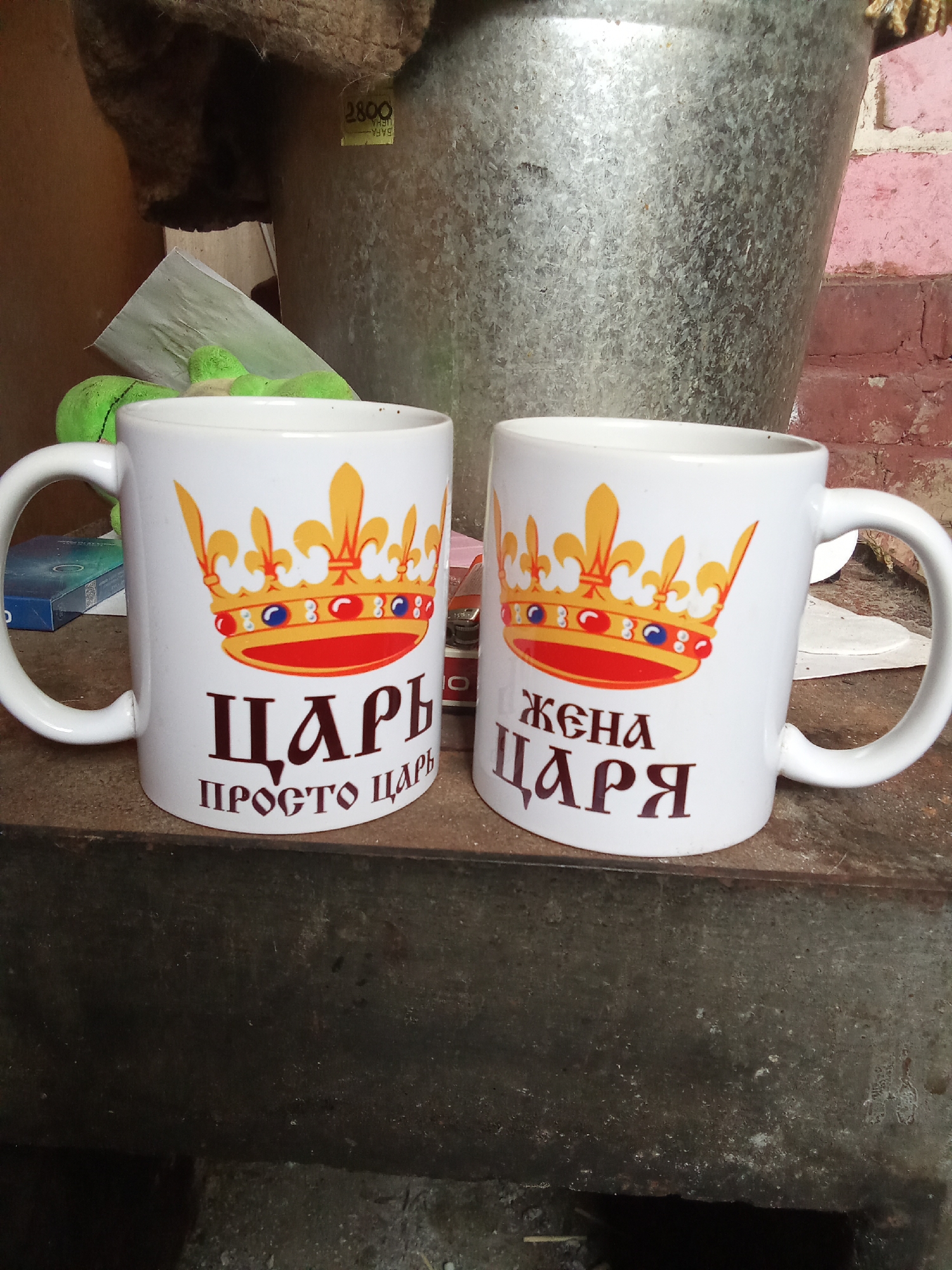 Two white ceramic mugs with crowns and Cyrillic text are displayed on a rustic metal surface. The mug on the left reads "ЦАРЬ ПРОСТО ЦАРЬ" (Tsar Just Tsar) and the mug on the right reads "ЖЕНА ЦАРЯ" (Tsar's Wife). The mugs have golden crowns with red and blue jewel accents. In the background, a large galvanized metal bucket and a brick wall are visible. The environment appears to be indoors or a sheltered outdoor area, with a hint of natural light. There are no people present in the image. The location is identified as Рощин ауылдық округі, Kazakhstan.