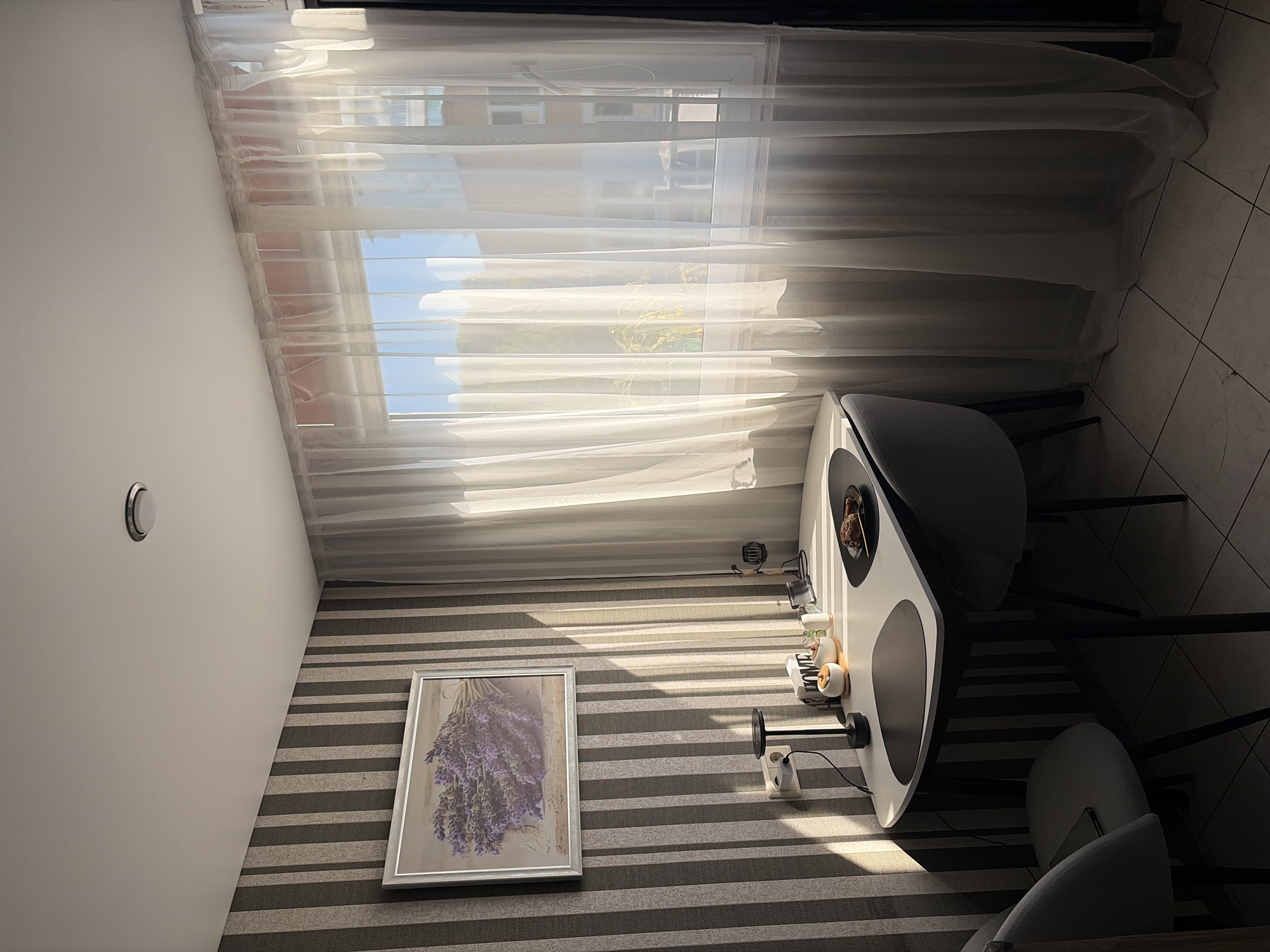 The image depicts a domestic interior scene, likely a dining area or a small kitchen nook, bathed in natural light. The room features a small, modern table with a white top and black legs, accompanied by two grey chairs. On the table, there are placemats, a small dish with food, and various decorative items including a jar with the word "HOME" and a small ceramic pot. A black desk lamp is also visible on the table, positioned near a power outlet on the wall. The wall behind the table is decorated with striped wallpaper in shades of grey and beige. A framed picture of lavender flowers hangs on the wall above the table. To the right, a large window is covered by sheer white curtains, allowing sunlight to filter into the room. The floor is tiled in a light beige checkered pattern. The overall impression is one of a well-lit, organized, and cozy living space. There are no individuals present in the scene. The image was taken inside an apartment in Sochi, Russia, as indicated by the lighting and architectural style which is common in residential buildings in the city.