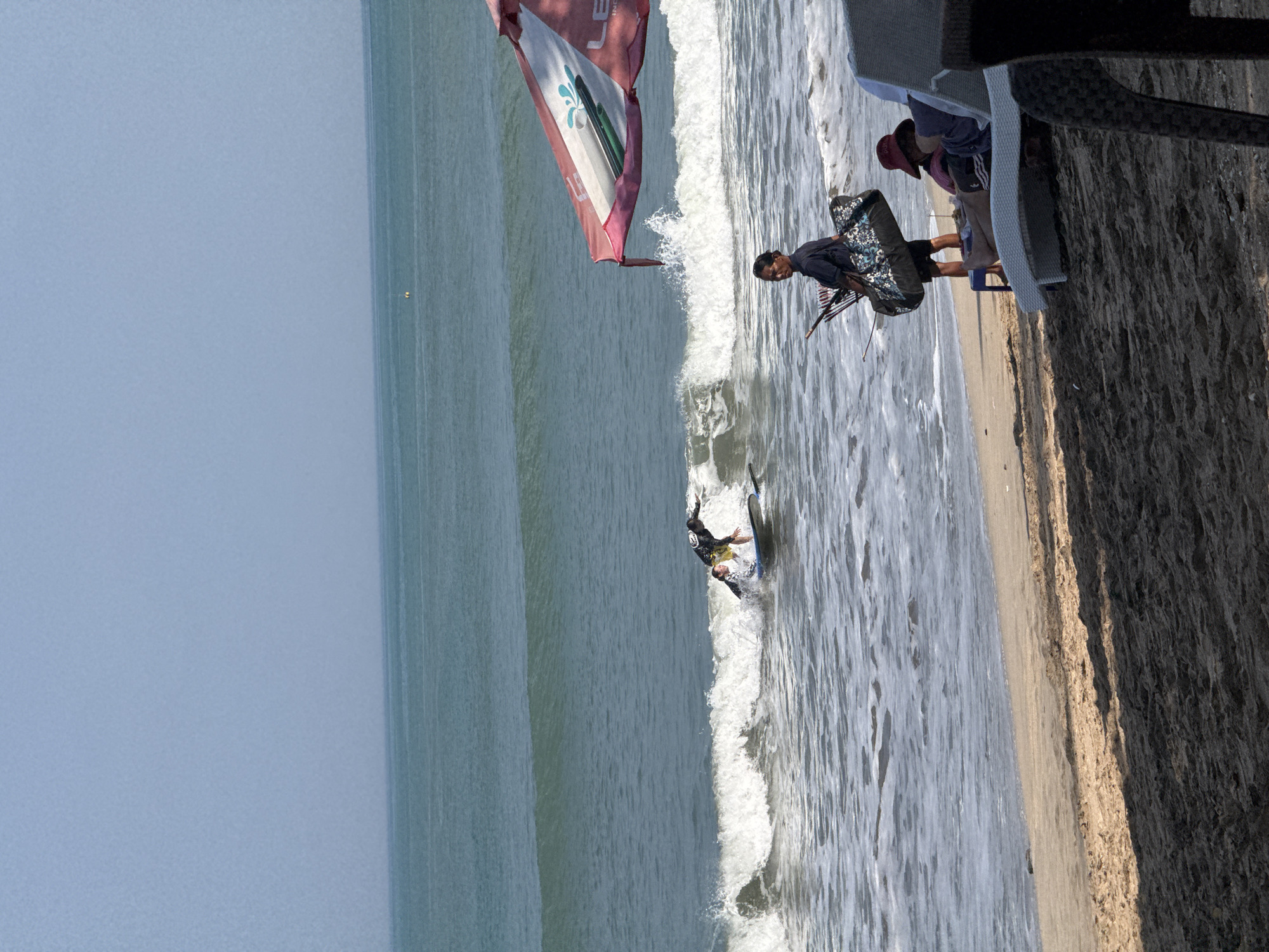 This image depicts a beach scene in Kuta, Indonesia. In the background, two individuals are surfing on a wave. The ocean is a turquoise blue, with white foam breaking on the shore. In the foreground, a man is walking along the beach carrying a large bag and fishing rods. To his right, a person is seated on a white lounge chair, wearing a red hat. A large pink and white umbrella is set up on the sand, partially obscuring the view of the surfers. The sand on the beach is light brown and appears wet near the waterline, with darker, dry sand further inland, casting shadows from unseen objects. The sky is a clear, pale blue.