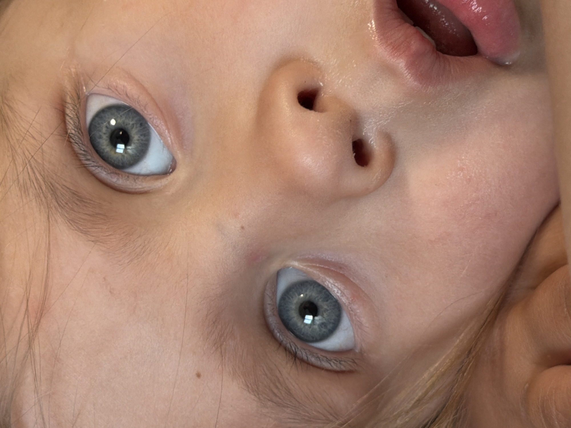 A close-up, angled view of a young child's face is presented. The child has fair skin and striking grey-blue eyes, with light eyelashes and eyebrows. The nose is small and upturned, with clearly visible nostrils. Part of the mouth is visible, revealing the upper lip and a glimpse of the tongue. The child's skin appears smooth and hydrated. Some strands of light-colored hair are visible near the edges of the face. The image does not provide any information about a location, city, or other aspects of the environment.
