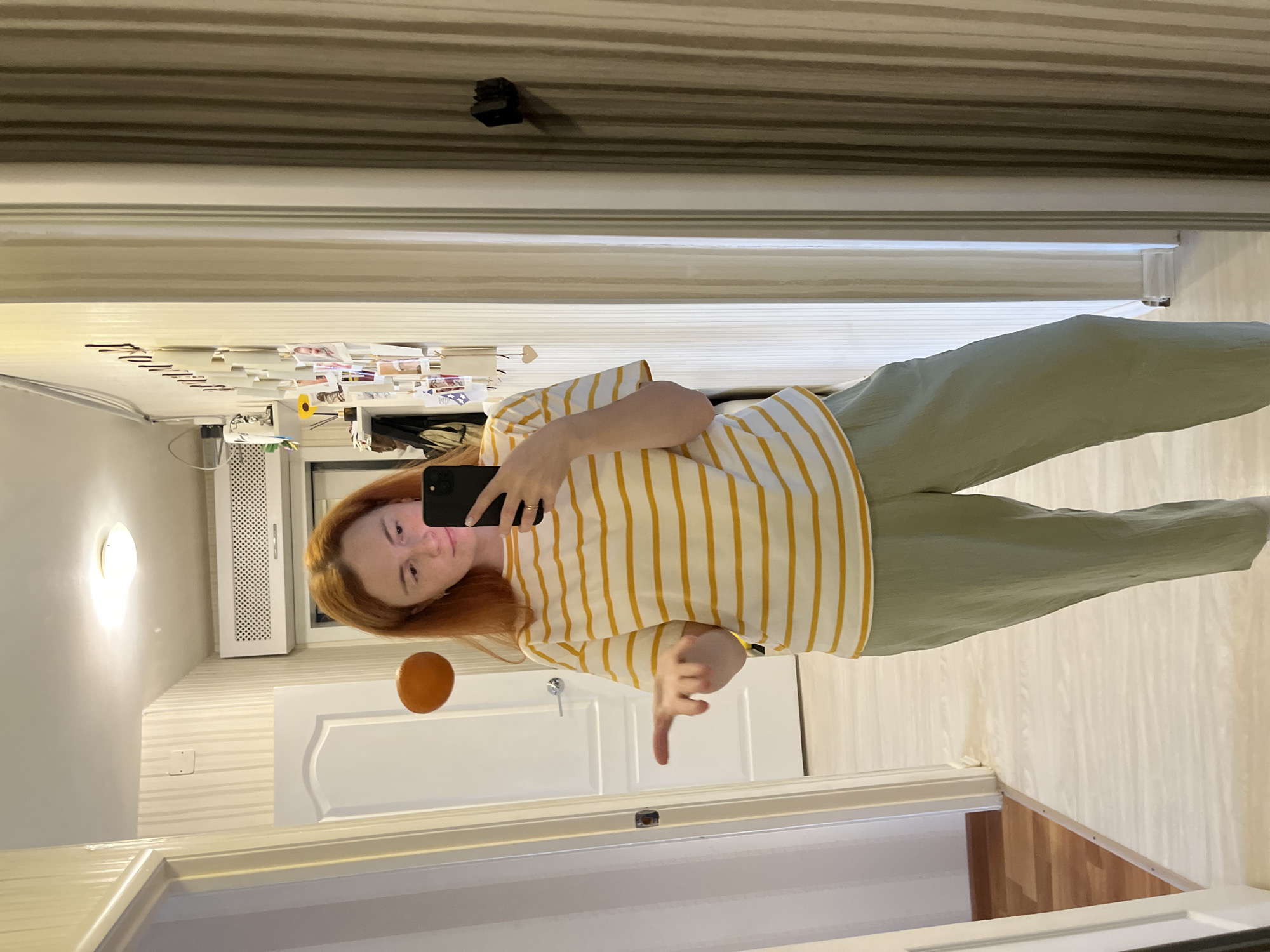 A young woman with red hair is taking a mirror selfie in a hallway. She is wearing a yellow and white striped t-shirt and light green pants. Her left arm is extended, with her hand open as if she is gesturing or explaining something. She is holding a black smartphone in her right hand. The hallway has light-colored walls with vertical striped wallpaper and a white door on the left. There is a round, orange object visible in the mirror's reflection to the left of the woman. The floor is light-colored with a wood-like pattern. The image appears to be taken indoors, likely in a residential setting. The location of Chaikovsky, Russia, is not visually identifiable from the image itself.