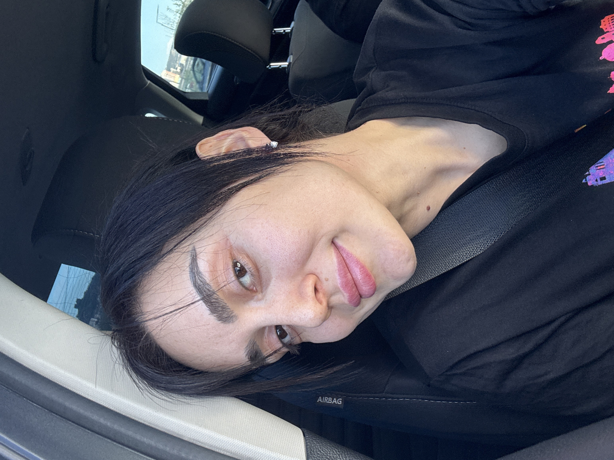 A person is seated in a car. They are wearing a black top and a seatbelt. The car interior is visible, including the seat, the door frame, and part of the dashboard. The location in the city of Tbilisi, Georgia is not discernible from the image.