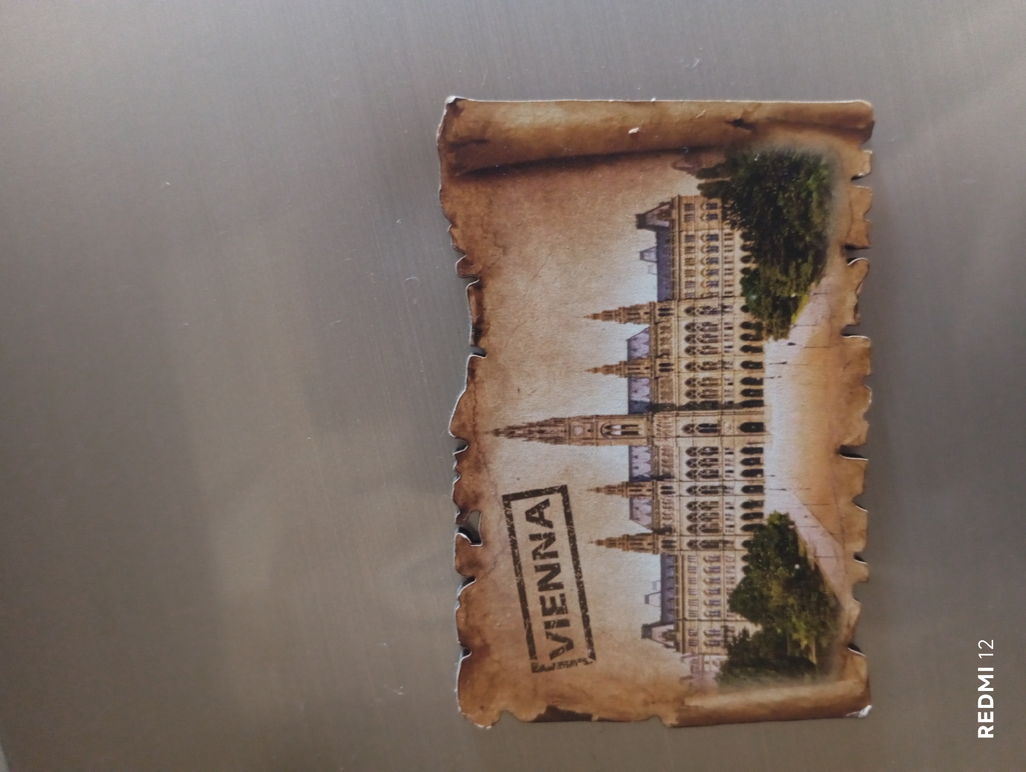 A magnet depicting the Vienna City Hall is attached to a metallic surface. The magnet is designed to look like a torn piece of aged parchment, with a stamped seal that reads "VIENNA" in a rectangular box. The City Hall is shown in the center of the magnet, with detailed architecture and trees in front of it. The metallic surface appears to be a refrigerator or similar appliance, with some faint scratches and reflections visible. The bottom left corner of the image shows the text "REDMI 12", indicating the device used to capture the photograph.