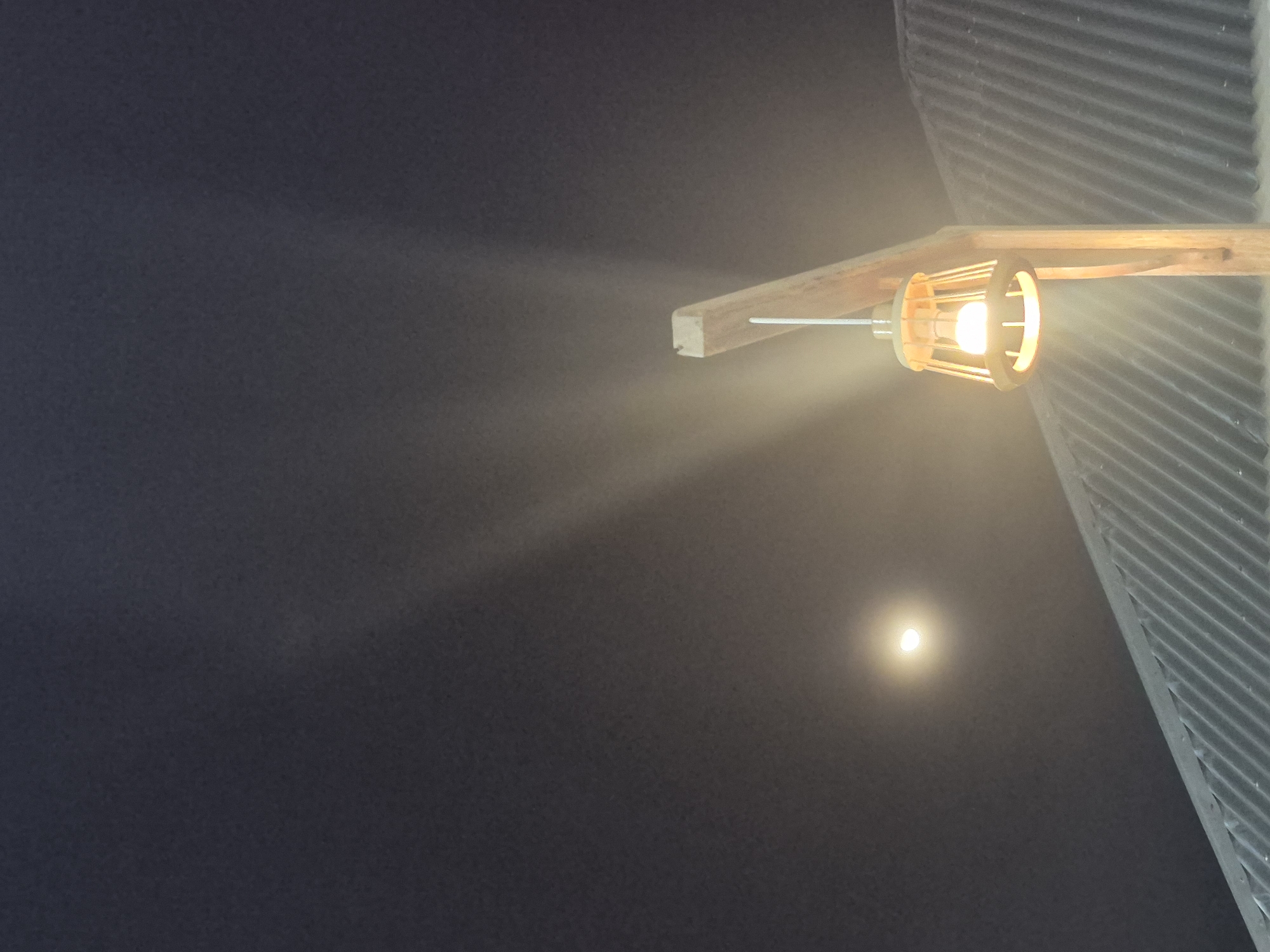 An upward-angled outdoor photograph taken at night shows a lighted bulb with a wood and metal cage fixture attached to a wooden support and a corrugated metal overhang. The bulb is illuminated, with a visible light stream. In the lower center of the dark background, a bright dot resembling the moon can be seen. The setting appears to be in Lhokseumawe, Indonesia, based on the provided context, but without more details, specific location or activity details are unidentifiable. There are no people visible in the photograph.