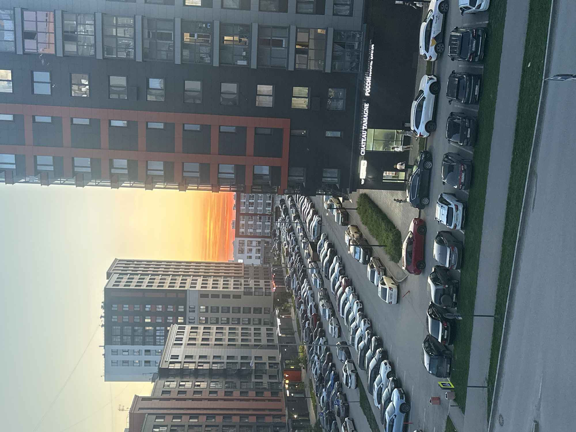 The image depicts a cityscape during sunset, likely taken from an elevated position. In the foreground, there is a large parking lot filled with numerous cars of varying colors and models. To the left, tall residential buildings dominate the scene, showcasing modern architectural designs with repetitive windows and some orange-colored accents. A sign reads "CHATEAU TAMAGNE" and Cyrillic script, indicating the location could be in Russia. A brief investigation identifies the location as Perm, Russia. The sky displays warm hues of orange and yellow, suggesting either sunrise or sunset. The overall scene conveys a sense of urban density and activity.