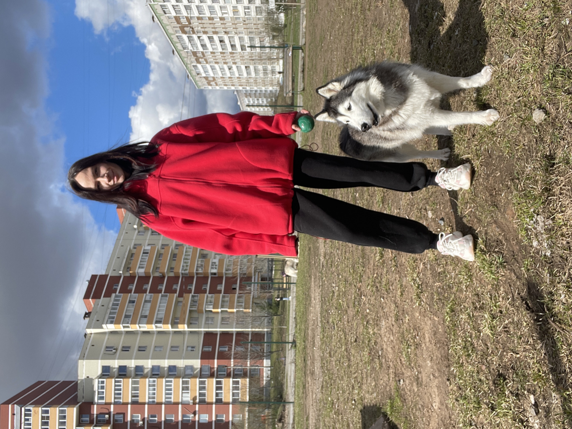 The image shows a woman standing on grass, holding a green ball in her hand, with a husky dog beside her, in Izhevsk, Russia. The woman is wearing a red jacket and black pants. The dog is lying on the grass. In the background are several apartment buildings. The sky is partly cloudy.