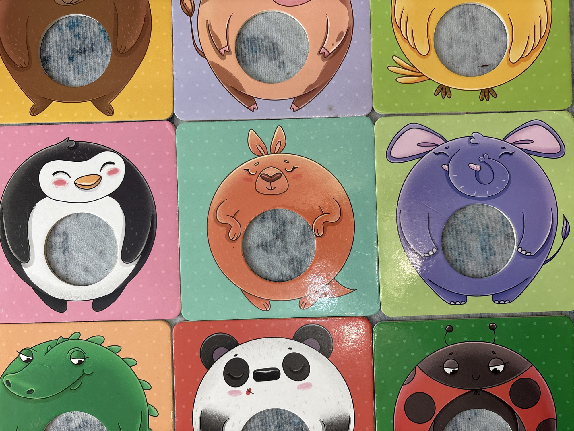 The image shows a collection of nine square cards with rounded corners. Each card features a cartoon animal character with a circular hole cut out in the center. The animals depicted are a bear, a cow, a bird, a penguin, a kangaroo, an elephant, an alligator, a panda, and a ladybug. The background colors of the cards vary and include yellow, lavender, green, pink, teal, purple, orange, red, and green again. The cards are arranged in a 3x3 grid. The location and relationship to the city of Izhevsk, Russia are unknown.