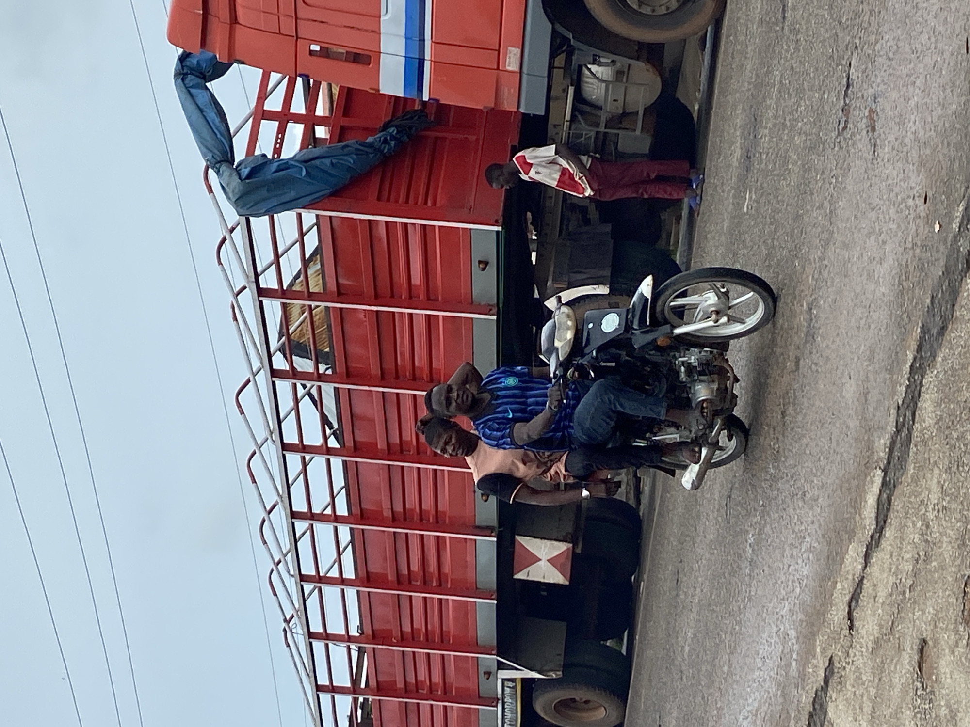 The image depicts an outdoor scene on a worn asphalt road in Mararaba, Nigeria, under an overcast sky with visible overhead power lines.
In the foreground, two men are riding a black motorcycle. The rider, a dark-complexioned male, wears a blue and black striped short-sleeved shirt and blue jeans, operating the motorcycle while looking towards the viewer and smiling. His left foot is visible in a sandal. Behind him, the male passenger, also dark-complexioned, wears a light brown and black short-sleeved shirt and dark pants, with his hair tied back. He is also looking towards the viewer and smiling, with his right arm resting on the rider's back.
To the left of the motorcycle, partially obscured by the vehicle, a third dark-complexioned male in a dark top is visible.
In the background, two large red trucks are present. The truck on the left is a flatbed, with high, slatted red sides, carrying what appears to be thatched material. A white and red "X" shaped hazard marker is affixed to its rear underside. The word "TOHORPOW" is partially visible in white text on its side. The truck on the right is the cab portion, also red, with a blue stripe detail. A blue tarpaulin is draped over the top of the rear truck's bed, extending towards the cab of the front truck. A fourth dark-complexioned male, wearing a white and red striped short-sleeved polo shirt and maroon trousers, stands barefoot on the roadside between the motorcycle and the front truck, leaning slightly against the truck's cab and looking downwards.
The road surface is uneven, showing cracks and patches of wear.