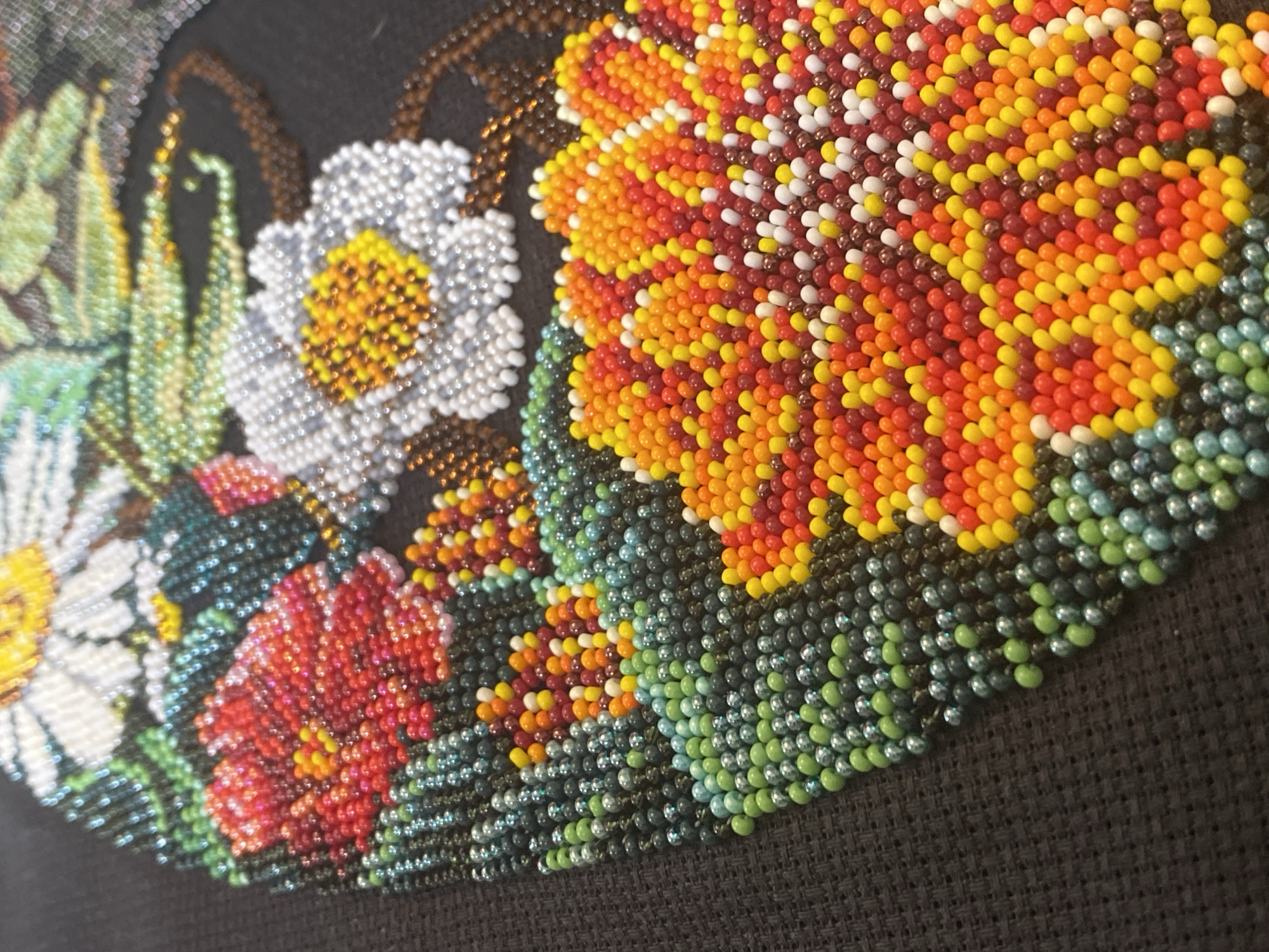 The image shows a close-up of a piece of beadwork depicting flowers and greenery against a black background. The beadwork appears to be a finished project, possibly an applique or panel. The flowers are rendered in various colors, including white, red, yellow, and orange. The greenery is in shades of green and blue-green. The beads are small and tightly packed, creating a mosaic-like effect. There is no indication of the location being Saint Petersburg, Russia.
