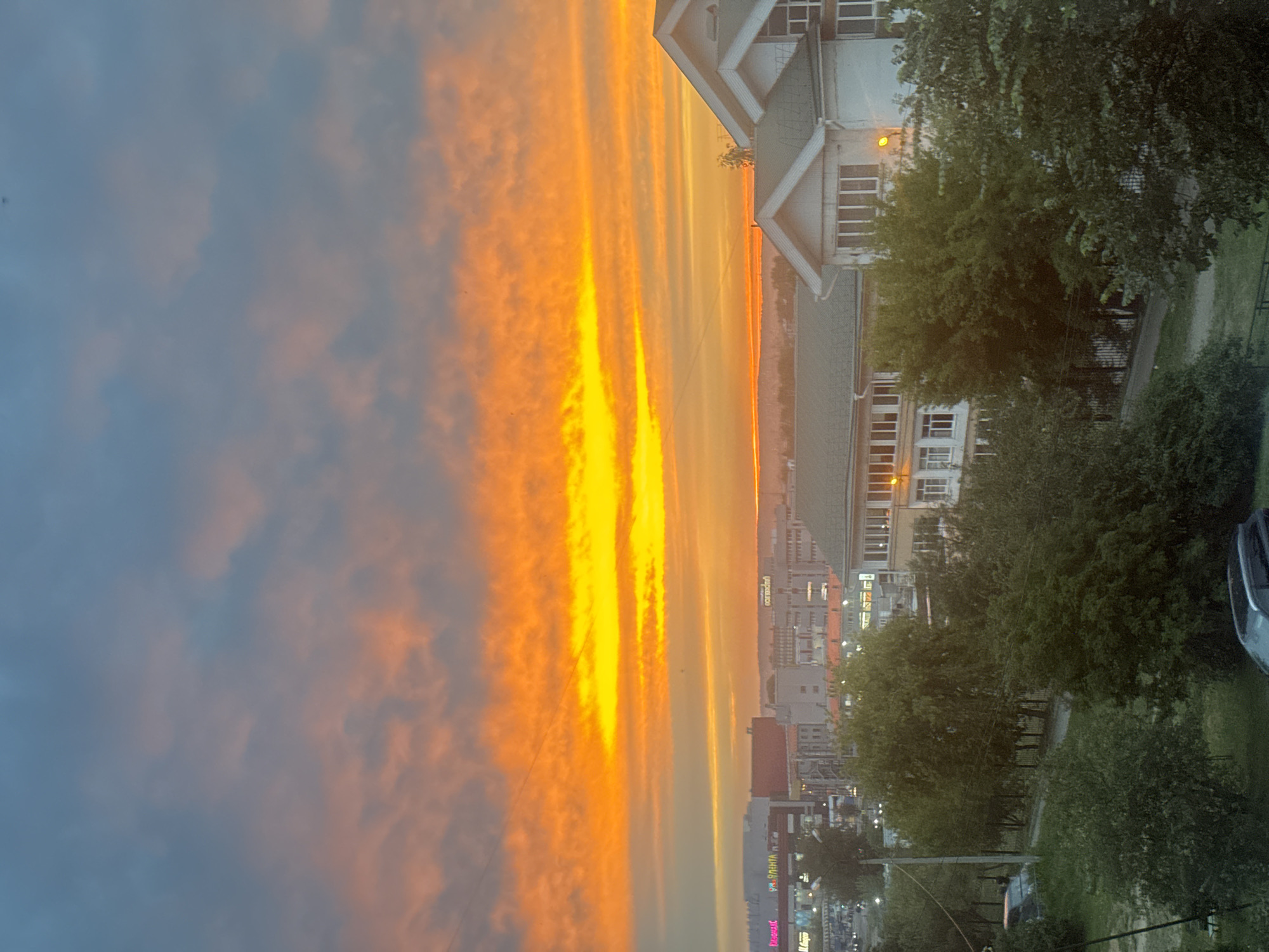 The image depicts a city skyline at sunset, characterized by a vibrant orange and yellow sky. The sun appears to have just set, casting a warm glow across the clouds. In the foreground, several buildings are visible, including what appears to be a multi-story residential building with a green roof and a larger commercial building with signage. Trees and greenery are scattered throughout the scene, suggesting a semi-urban or suburban environment. The sky is partly cloudy, with streaks of orange and yellow clouds dominating the upper portion of the image. The lower portion shows buildings and trees, with hints of a road or pathway. The image seems to be taken from a higher vantage point, looking across the city. Based on visual cues such as architectural styles and the presence of Cyrillic signage on some of the buildings, the location can be identified as Irkutsk, Russia. Specifically, one of the buildings has visible signage including "ЭЛЕНТА" and "ОКЕЙ", which are consistent with businesses found in Russian cities. The overall scene captures the tranquil beauty of a sunset over an urban landscape.