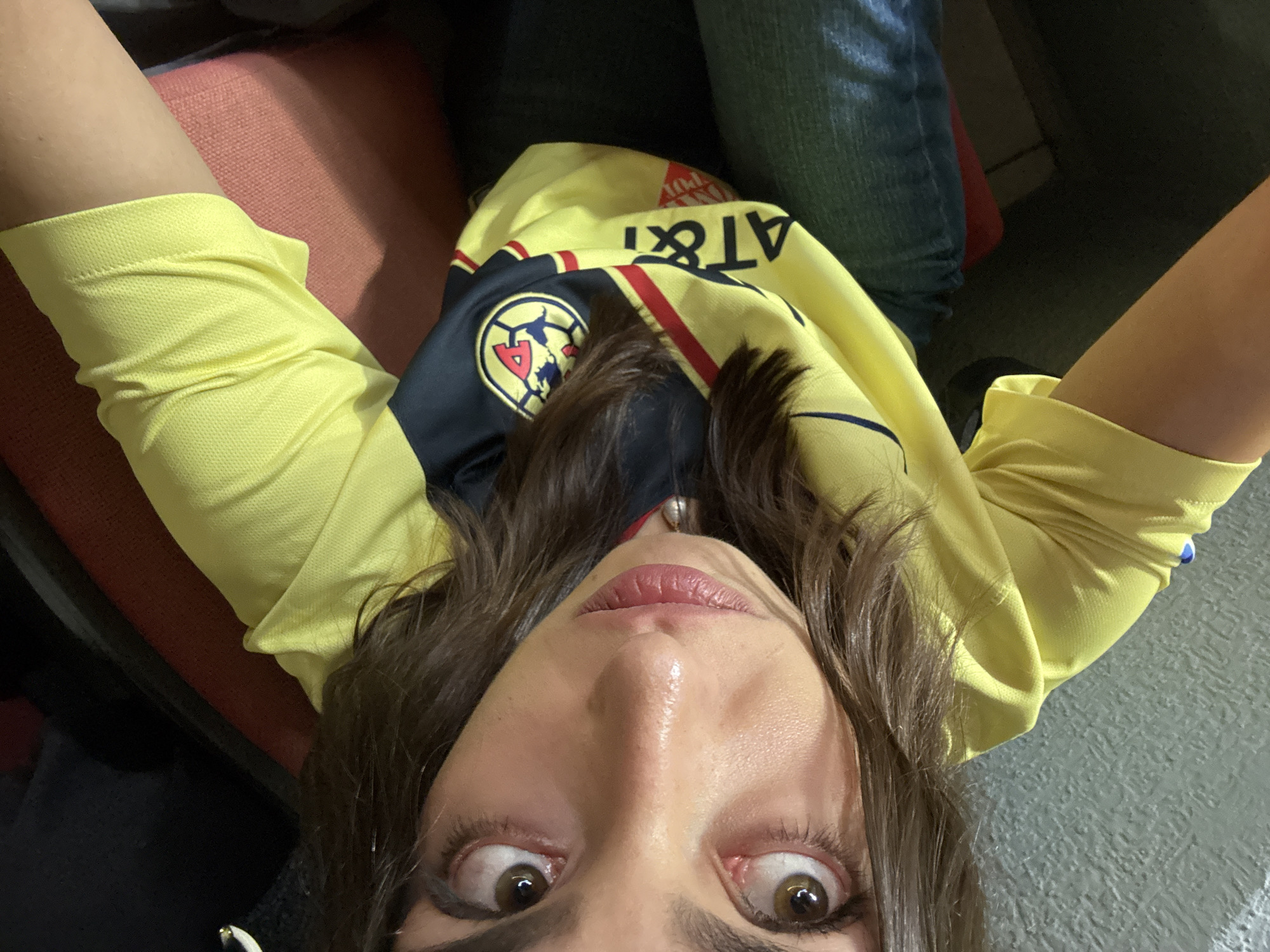 A woman is taking a selfie, wearing a yellow and black Club América soccer jersey with "AT&T" printed on it. She is seated, wearing jeans, possibly in a stadium or public seating area. Her expression shows exaggerated surprise. The location appears to be indoors, though specific details about Mexico City are not discernible from the image.