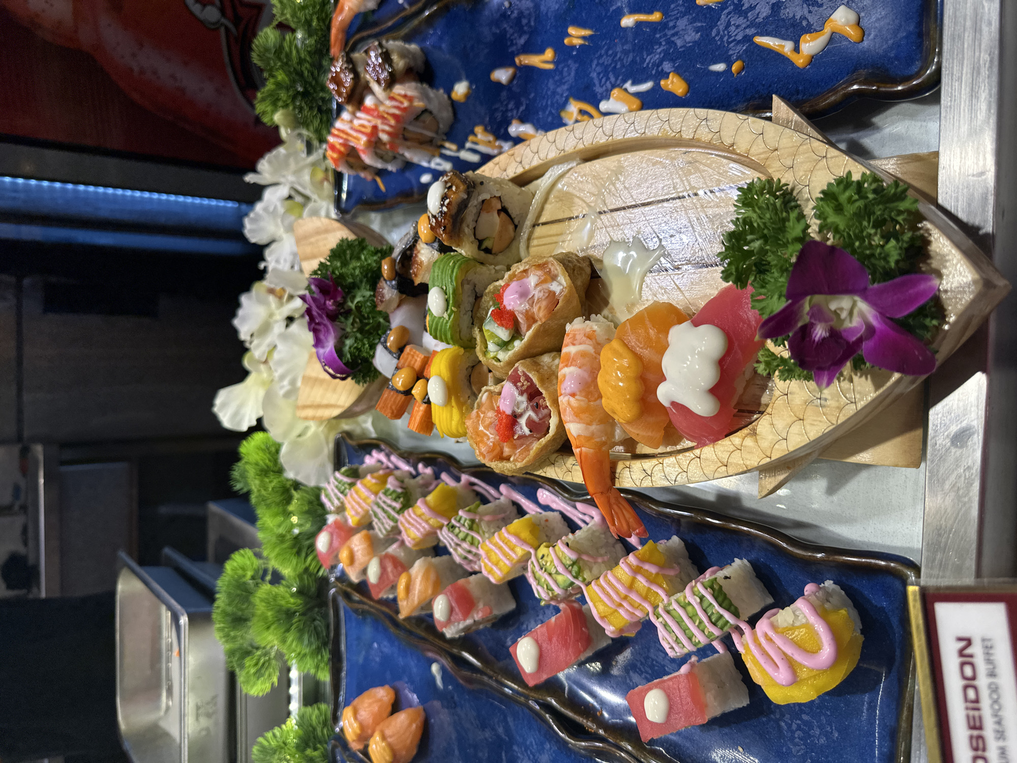 The image displays an elaborate and colorful assortment of sushi and sashimi arranged on decorative platters, likely part of a buffet spread. The food is presented with attention to detail and aesthetic appeal.
In the foreground, a blue ceramic platter with wavy edges holds rows of various sushi rolls and nigiri. These include pieces topped with red tuna, pink-drizzled rolls, yellow mango slices, and green avocado slices. Some pieces feature a contrasting white or pink sauce drizzle on top. Alongside these are individual pieces of what appear to be salmon roe or similar small orange seafood.
Above this, a wooden, fish-shaped boat platter showcases a different selection of sushi and sashimi. This includes shrimp nigiri, tuna nigiri, inari sushi (rice in fried tofu pockets), and various sushi rolls with fillings like eel, crab stick, and fish. Some pieces are garnished with orange and white sauces. This platter is adorned with fresh-looking green parsley-like garnishes and vibrant purple and white orchids, enhancing the visual appeal. Further up, another blue platter holds more sushi rolls, also decorated with orange and white sauces.
The background suggests an indoor setting, possibly a restaurant or hotel buffet, with metal food containers visible on the left and a red banner or sign in the upper left. The overall arrangement indicates a professional food service environment.
A red-brown sign is visible in the bottom right corner, partially revealing the text "POSEIDON" and "MIUM SEAFOOD BUFFET," suggesting the context of a seafood buffet. The lighting is bright, highlighting the colors and textures of the food.