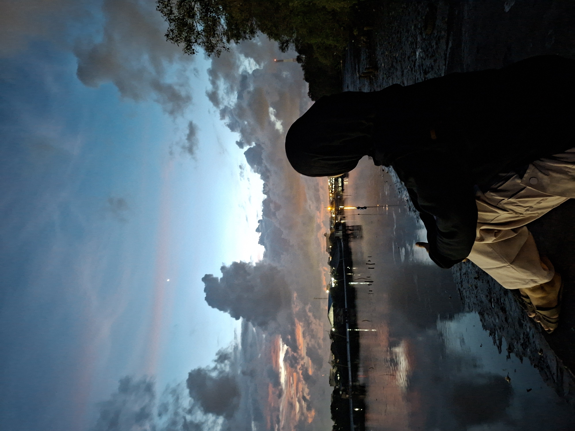 The photograph captures a serene landscape near a body of water at dusk, likely within the city of Lhokseumawe, Indonesia. The sky is a canvas of deep blues and grays, with clouds reflecting the fading light. A long strip of orange-tinged sky hints at the setting sun. A person stands on the edge, their form cloaked in a dark hooded jacket and light-colored pants, possibly contemplating the scene. The person is looking towards the water. The water reflects the skyline of the city, indicating the presence of buildings and lights in the distance. The foreground shows the edge of the water and the shoreline.
