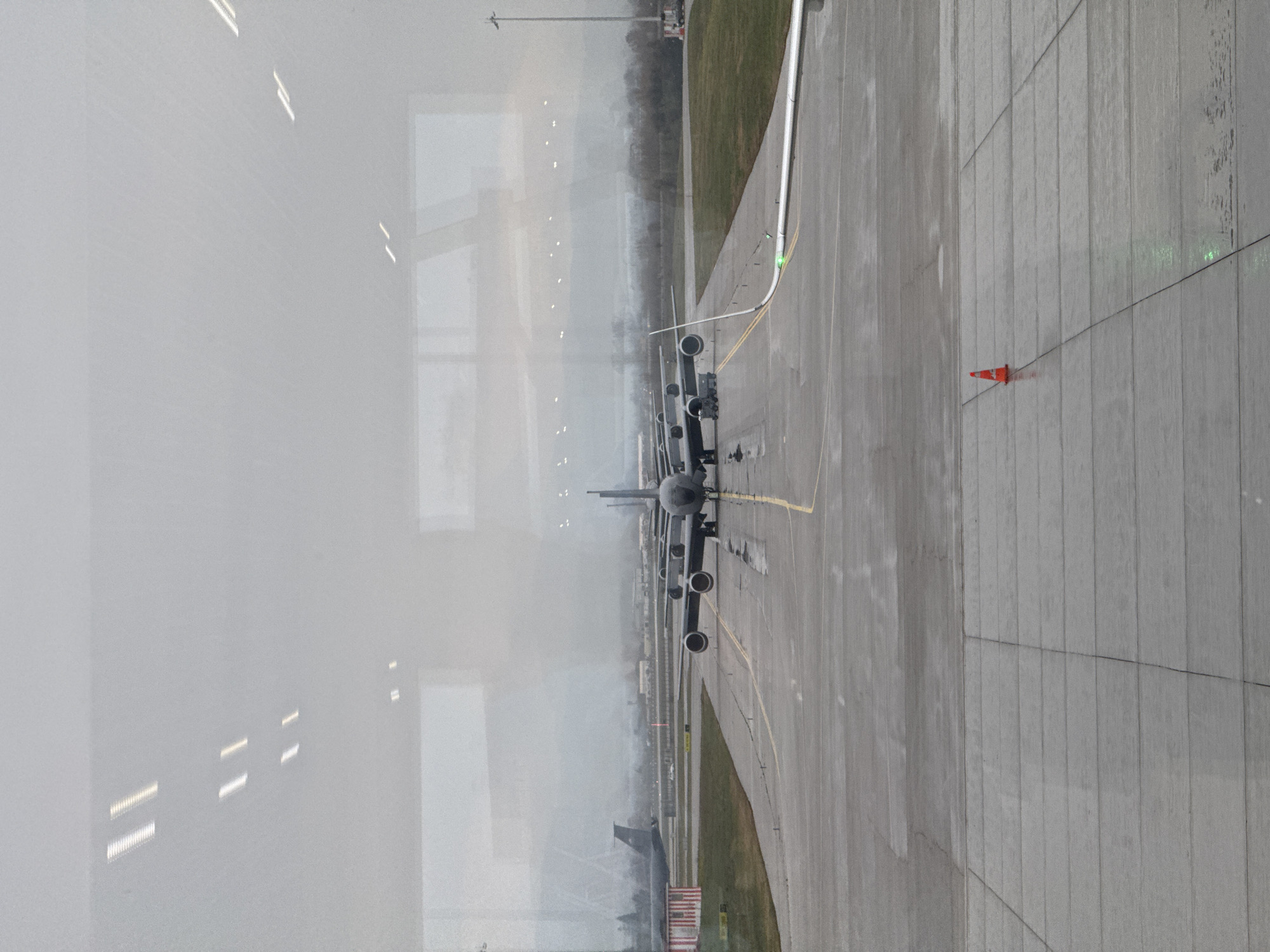 The image shows a view of an airport tarmac, likely viewed through a window. Several airplanes are parked on the tarmac, lined up one behind the other. An orange traffic cone is visible on the ground near the edge of the tarmac. The weather appears overcast or foggy, with a low-visibility horizon. The location is likely at the Sofia Airport, Bulgaria, given the context. There are no people visible in the image. The planes are stationary, with no apparent activity around them.