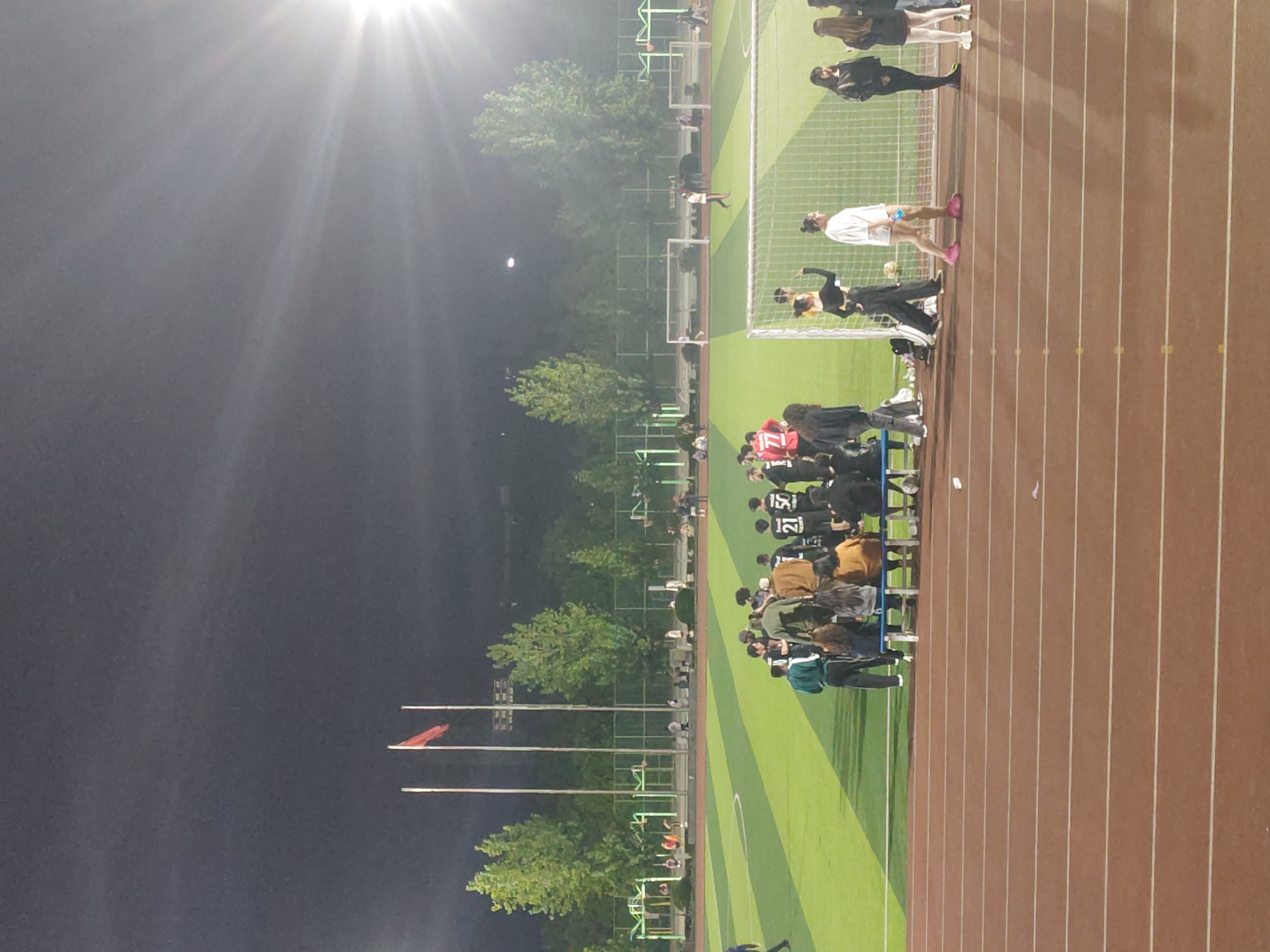 This image captures a nighttime scene at an outdoor sports complex, likely in the Chongqing High-tech Zone, China. The primary features are an artificial turf soccer field with goalposts and a multi-lane running track surrounding it. The field is brightly illuminated by floodlights, creating a strong lens flare effect in the upper left quadrant, and the dark sky reveals a distinct moon.
Numerous people, primarily young adults, are gathered around the field. A large group stands and sits along the sideline and on the running track, suggesting spectators or team members awaiting their turn. Several individuals wear sports jerseys with numbers visible, such as "11", "21", and "15". One person in a white shirt and pink athletic shoes stands on the soccer field near a goalpost.
The presence of players in jerseys, goalposts, and a large gathering indicates a sporting event, possibly a soccer match or practice session, taking place under artificial light. Trees line the perimeter of the facility, and several tall flagpoles, one displaying a red flag, are visible on the left side of the frame. The scene conveys an active evening at a community or school sports venue.