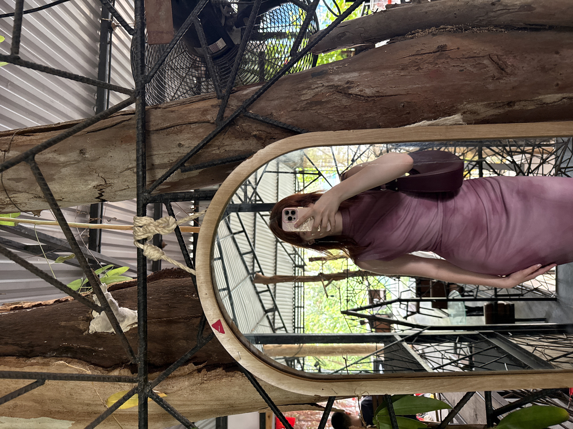 The image, rotated 90 degrees counter-clockwise, depicts a mirror selfie taken by a woman within a distinctive indoor or semi-outdoor setting in Bac Nha Trang Ward, Vietnam.
The central subject is a woman, reflected in an oval mirror with a light wooden frame. She has reddish-brown hair and wears a fitted, light purple or mauve dress. Her face is mostly obscured by a light pink smartphone, which she holds up to take the photograph. A dark, possibly burgundy, handbag is slung over her shoulder. Her hand is visible resting on her hip.
The setting visible outside the mirror's reflection incorporates a blend of natural and industrial elements. Large, rough wooden logs or tree trunks are integrated into an elaborate dark metal framework or trellis that spans across the scene. A large, dark industrial fan is positioned within this metal structure at the top. Behind these, a corrugated metal surface, possibly a roof or wall, is visible. Green foliage and small plants peek through the metalwork, adding natural accents. A piece of braided rope is tied around one of the wooden logs.
Within the mirror's reflection, the background offers a wider view of the establishment's design, revealing more of the metal framework, corrugated structures, and abundant green trees, suggesting an open or naturally lit space. It is daytime, indicated by the bright, natural light. A small red triangular sticker is visible on the bottom left edge of the mirror's wooden frame. The smartphone held by the woman clearly displays "IPHONE" branding on its back. The wooden elements show natural cracks and textured bark.