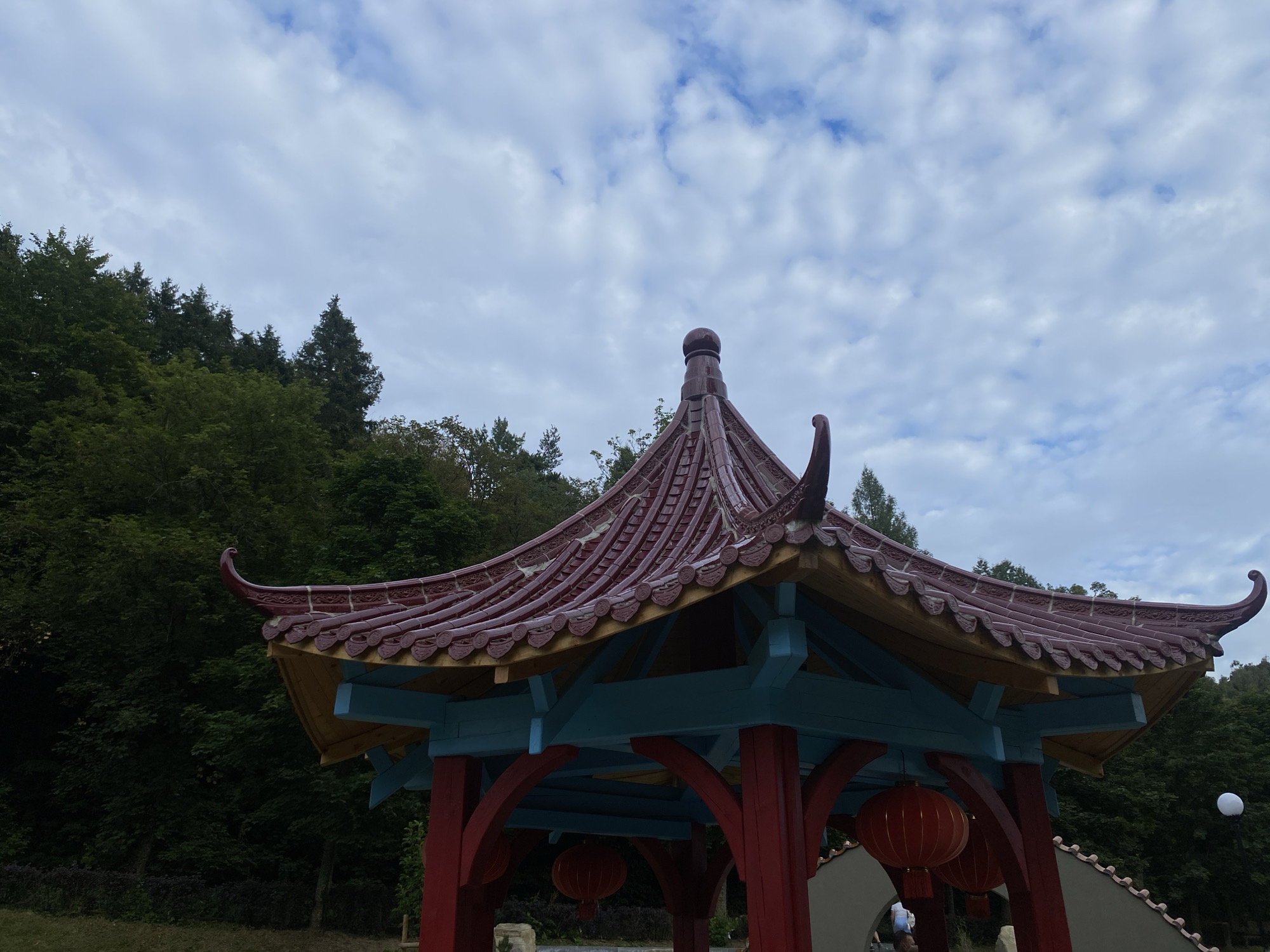 The image shows a traditional Chinese-style pavilion with a red tiled roof and red and blue painted wooden supports. Two red paper lanterns hang from the pavilion's interior. The pavilion is situated outdoors, with a dense green forest visible in the background. The sky is cloudy and light blue. The scene is located in Muszyna, Poland, at the Mountain Garden (Ogród Sensoryczny) which features themed areas, including one with Asian influences, as indicated by the pavilion's architecture.