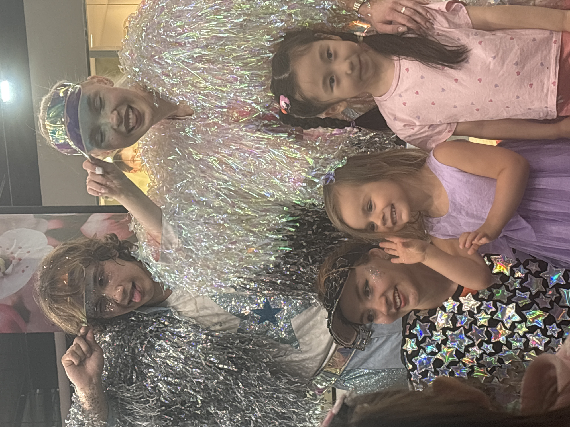 The image shows a group of people, including children and adults, posing for a photograph indoors. The setting appears to be a festive event or party. The adults and one child have makeup on, and they are wearing shiny, star-themed, festive costumes. The other children wear pink and purple dresses. The location, city, or specific scene details are not discernible from this single image.