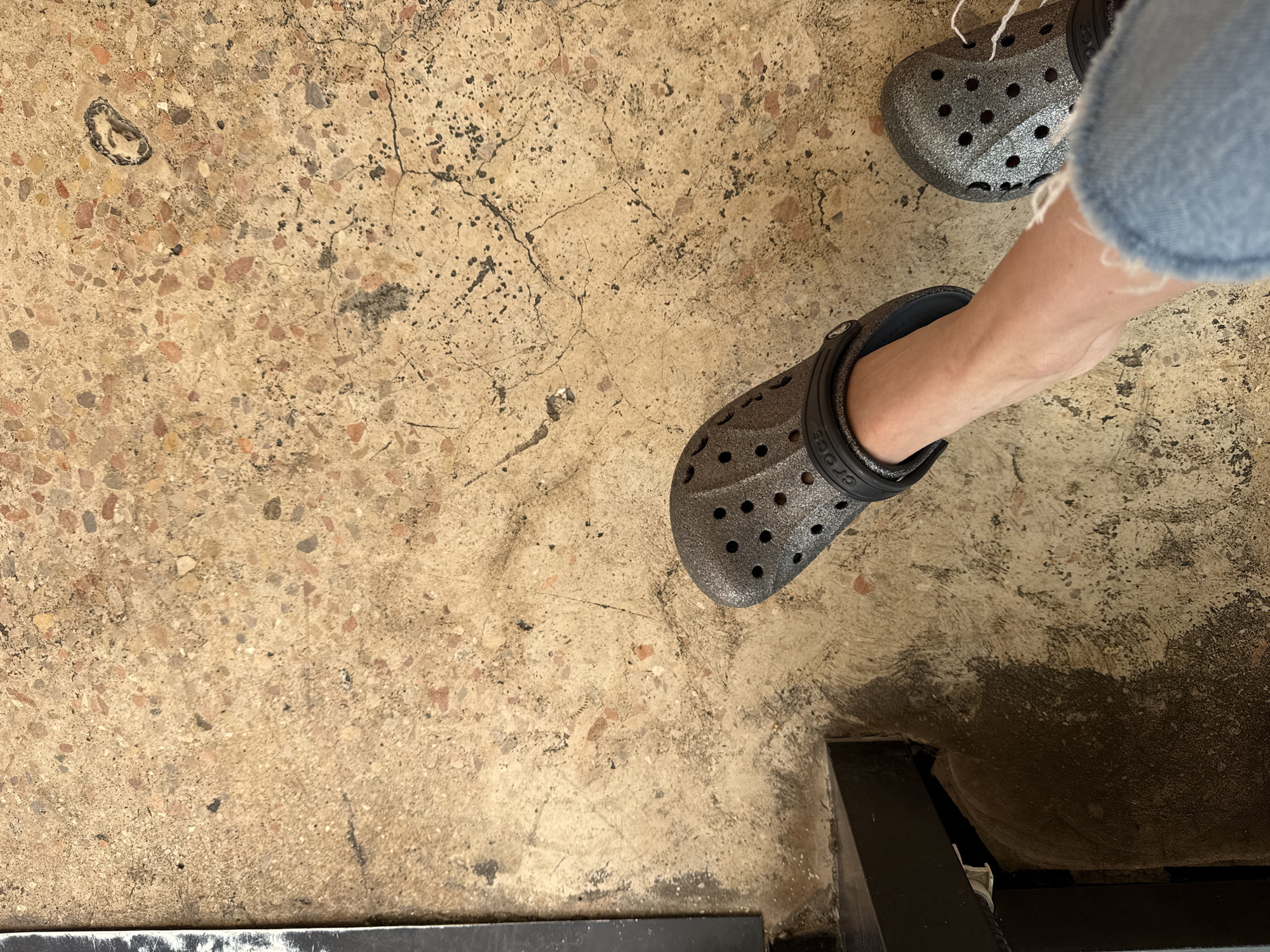 The image shows a pair of silver Crocs, one on a person's foot, on a terrazzo-style floor with cracks and imperfections. The lower part of a jean-clad leg is visible. A dark, possibly metal, object or fixture is partially visible in the bottom right corner of the image, and a white object is partially visible in the bottom left corner. The background is a concrete floor. It's impossible to determine whether this image was taken in Tel-Aviv, Israel without further evidence.