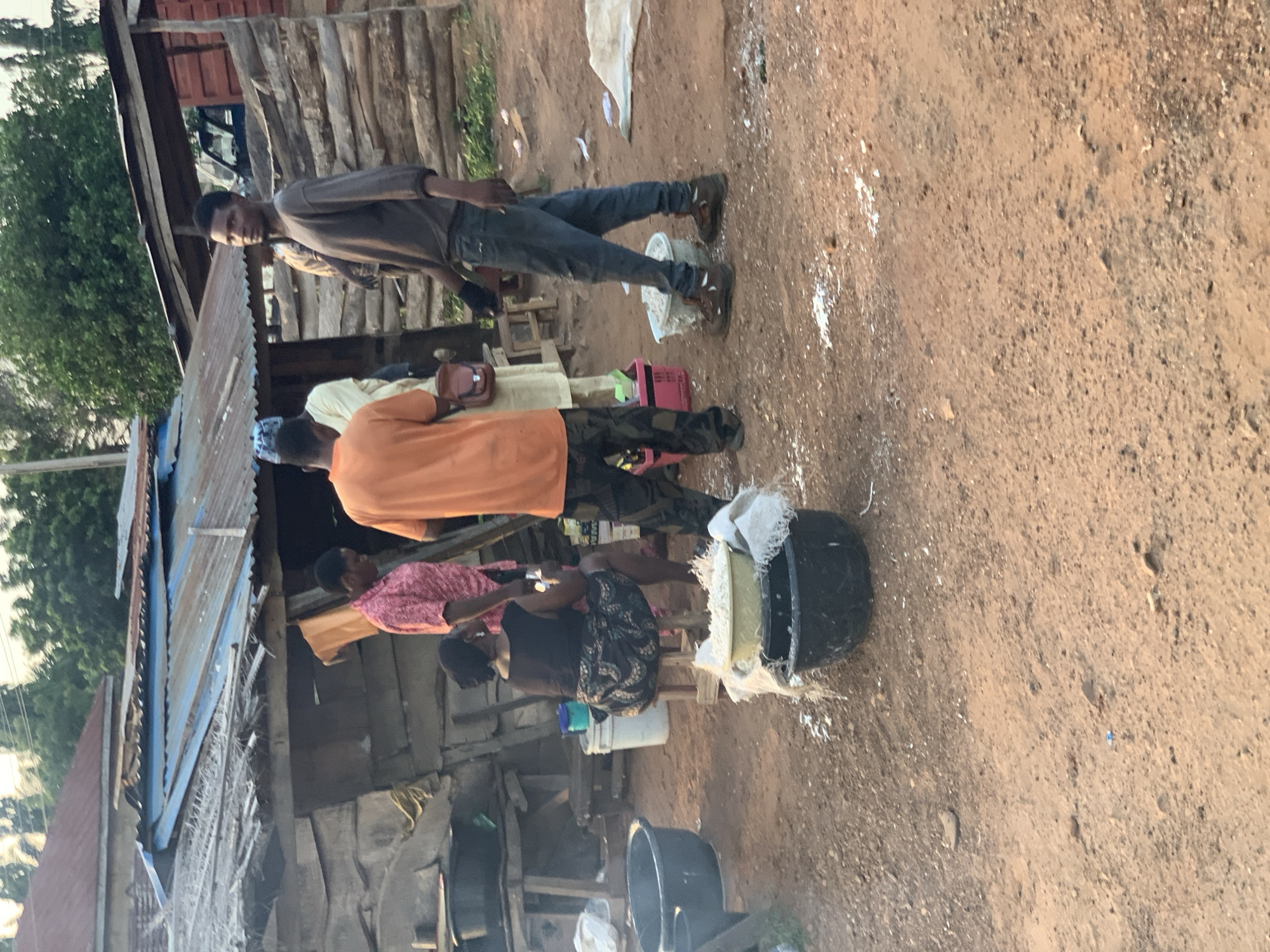 The image depicts an outdoor scene in Gbeleko, Nigeria. Four people are visible, positioned near a wooden structure with a corrugated metal roof.
One individual, wearing a brown shirt and jeans, stands facing the camera. Another person in an orange shirt and camouflage pants is standing near a third individual wearing a pink shirt. A fourth person, in a dark shirt with a patterned skirt, is seated. A black bucket filled with a white substance sits nearby.
The ground is dirt, with scattered white matter around the bucket. The background includes a green foliage and wooden structures.
