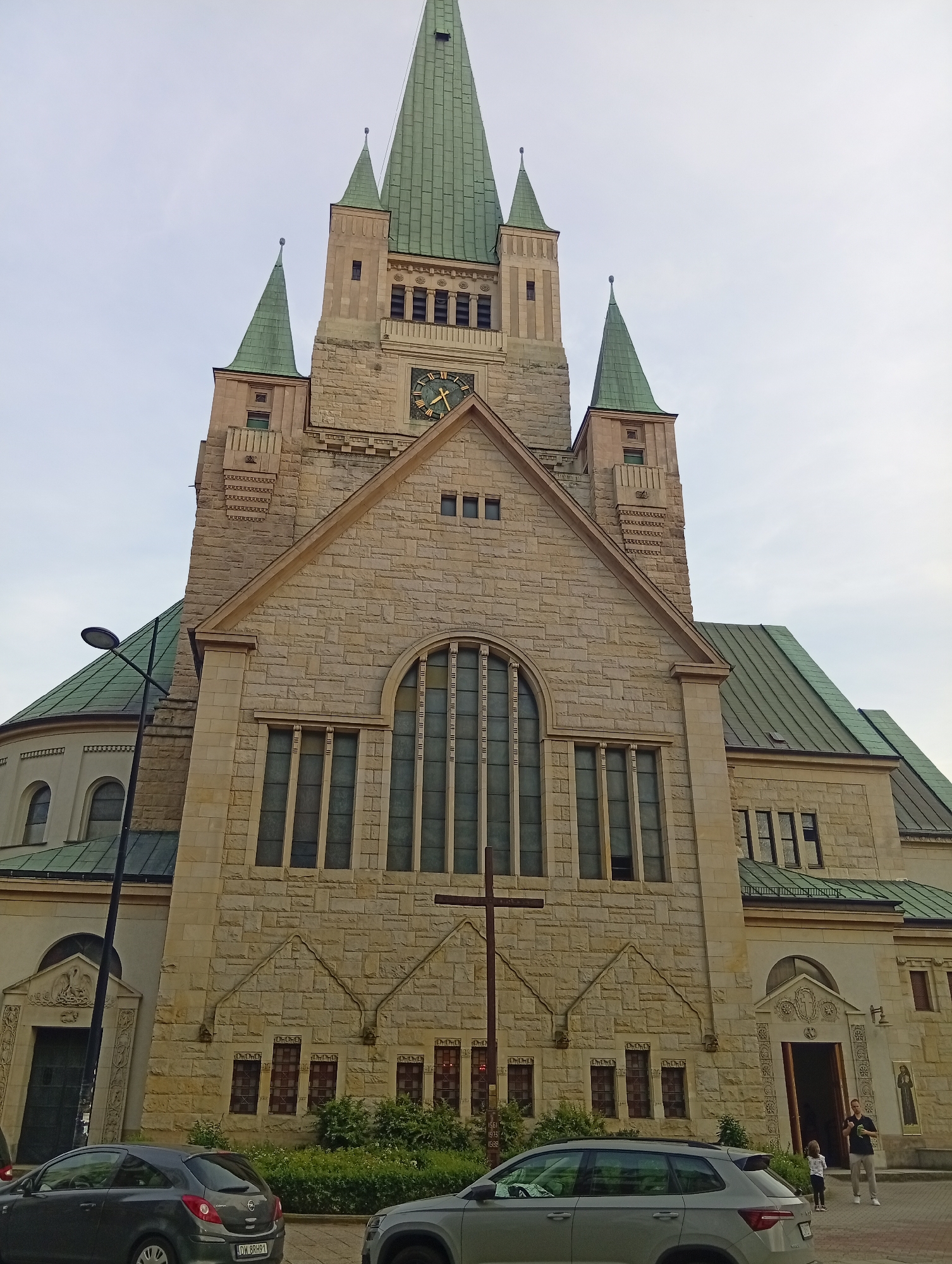 The image shows a frontal view of the Neo-Romanesque Basilica of the Sacred Heart of Jesus in Wrocław, Poland, under a cloudy sky. The church's facade is constructed of light-colored stone blocks, with a prominent central tower featuring a clock and topped by a green copper spire. Two smaller towers with similar green spires flank the central one. Below the clock, a large, arched window with multiple panes dominates the upper part of the facade. A large wooden cross stands in front of the church. The lower part of the building has several smaller windows and two entrances, one of which is open. Green bushes and trees line the front of the church. Two cars are parked in front of the church, and two people, a man and a child, are standing near the open entrance. The architecture is detailed and imposing, with ornamental carvings visible around the entrances and windows.