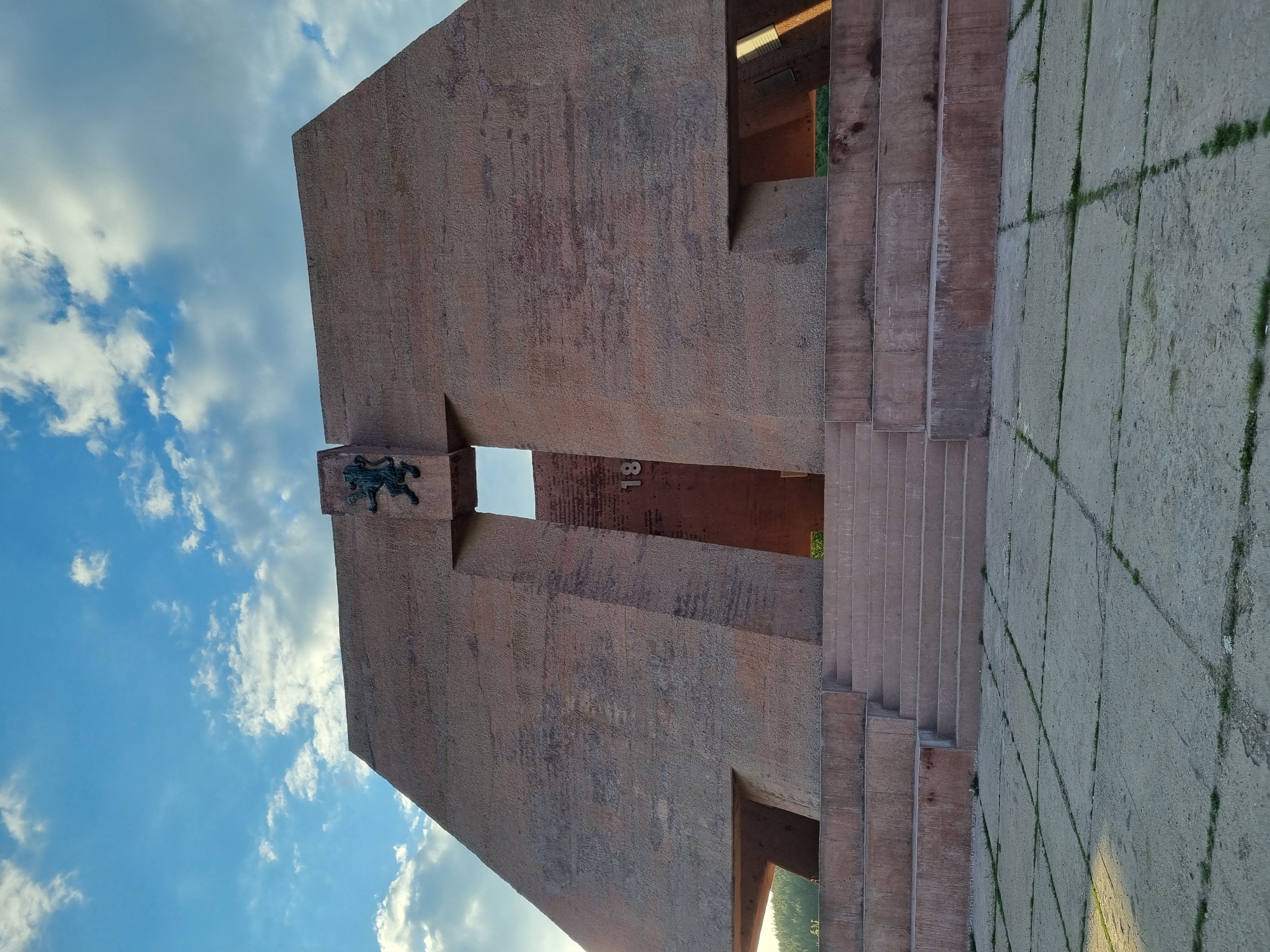 The image shows the Monument "Chavdar" in Gurgulyat, Bulgaria, with its monumental concrete structure, a relief depicting a lion, and visible steps leading up the side. The sky is partly cloudy.